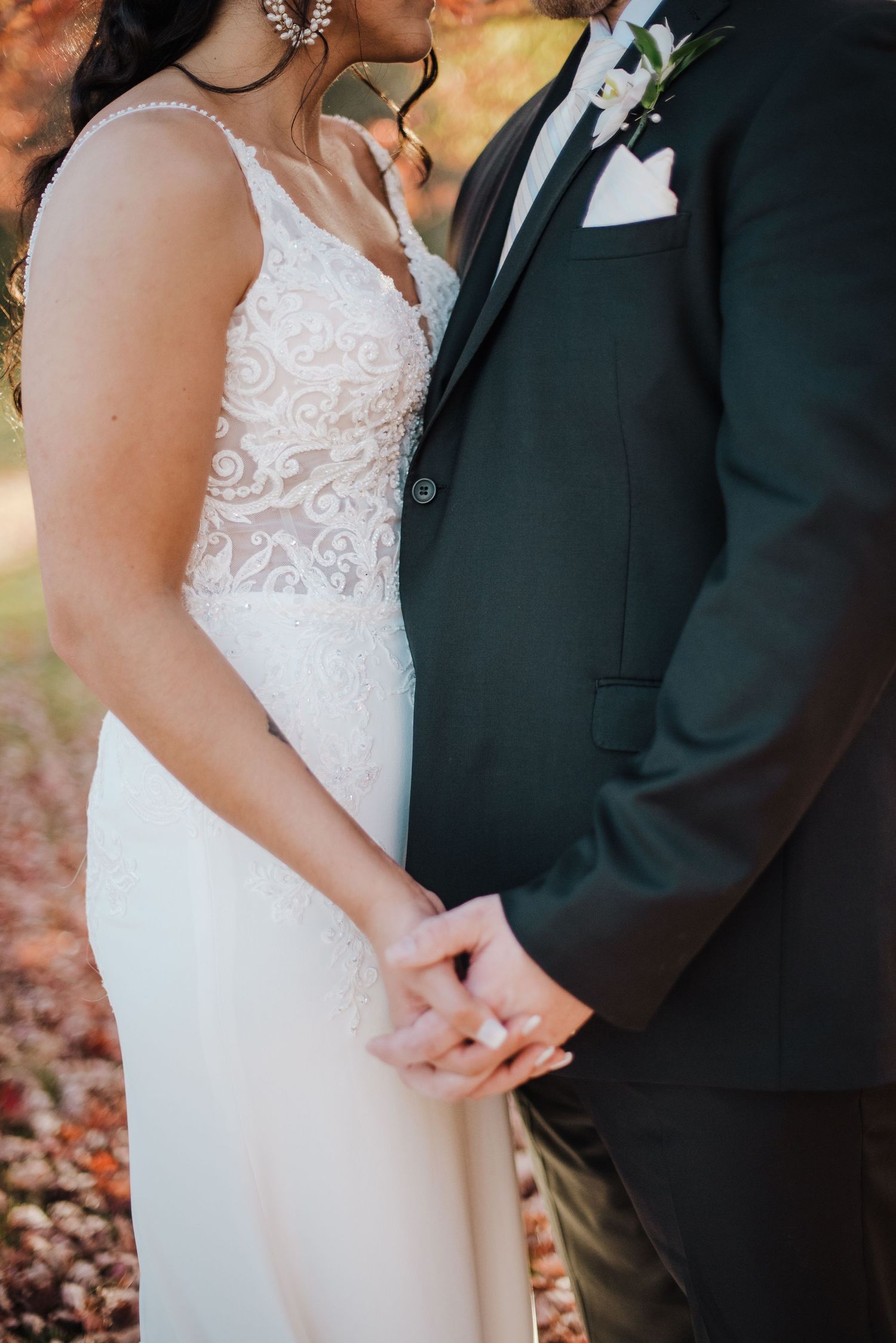 Close-up of bride in lace bodice gown and groom in black suit holding hands tenderly with autumn foliage background.