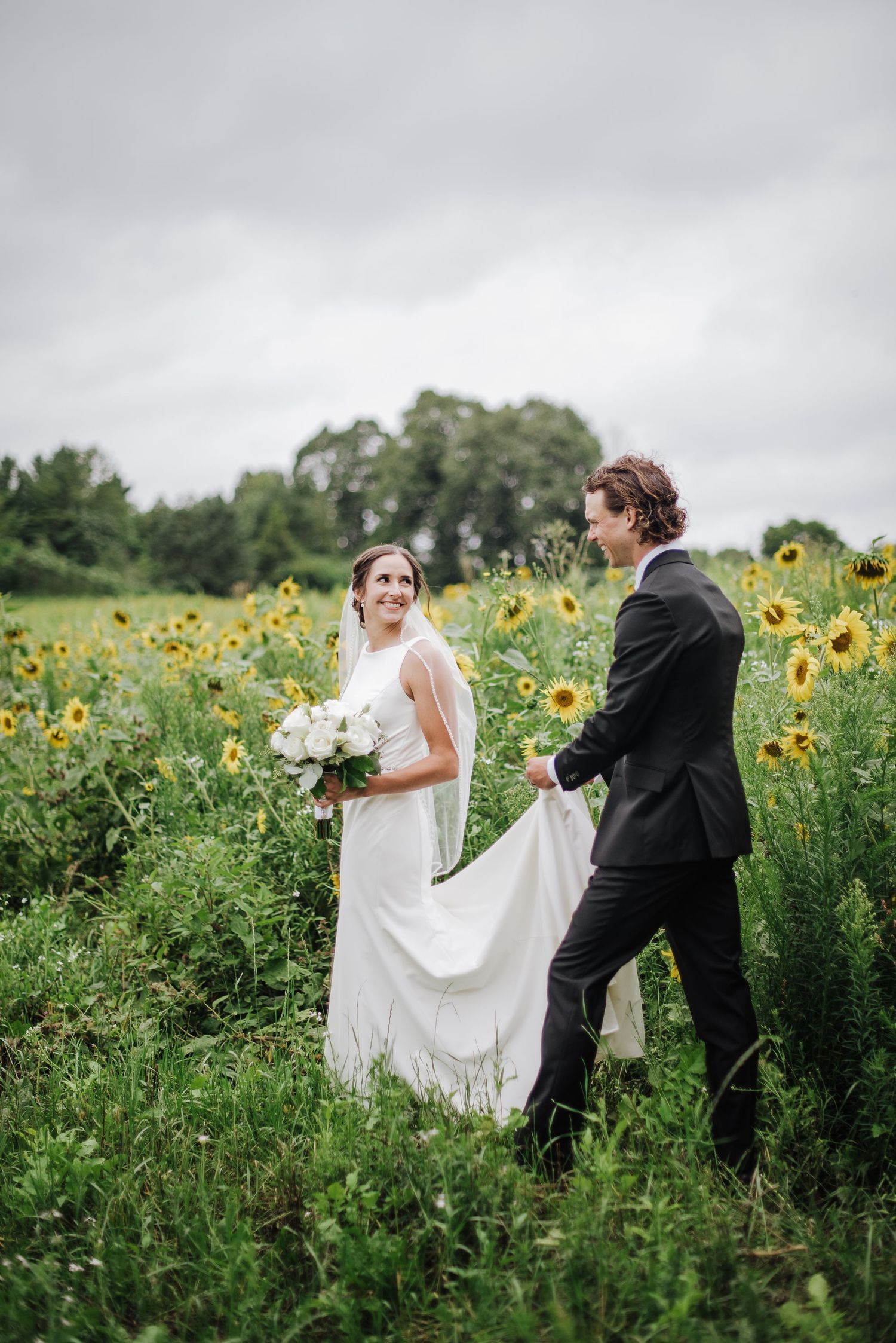 Groom carries bride's train as she walks smiling through a lush sunflower field on a cloudy day.