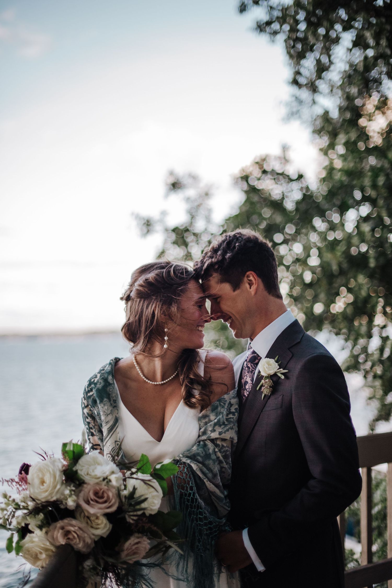 Bride and groom touching foreheads romantically near lakeside at dusk, bride holding white rose bouquet wearing green wrap.