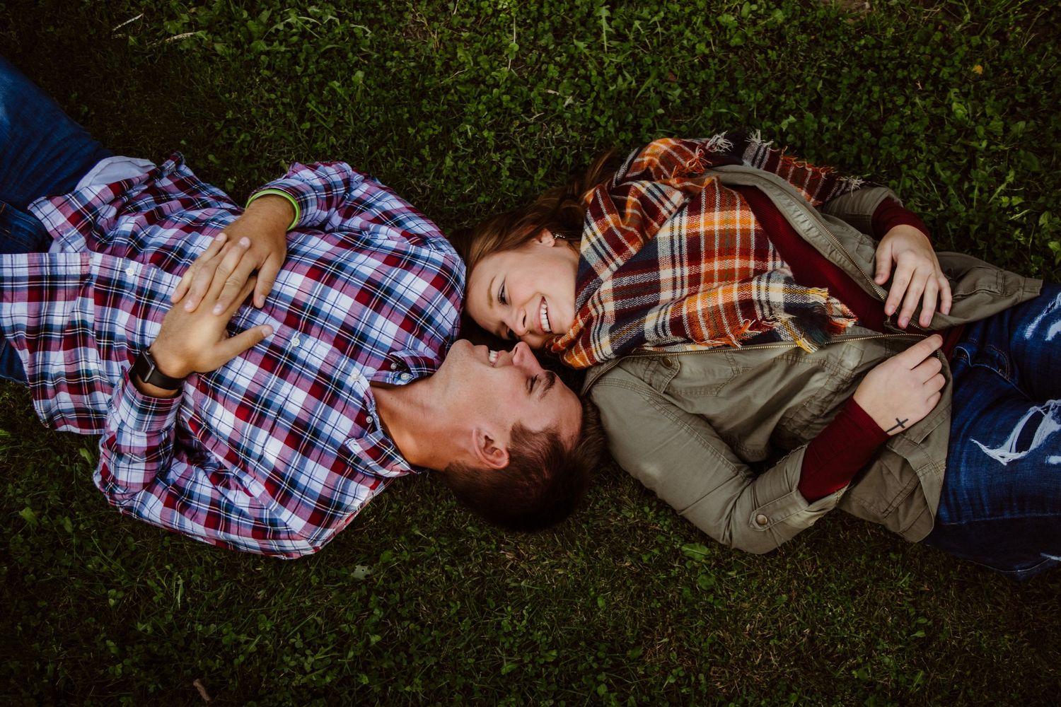Couple lies on green grass looking at each other lovingly in a casual outdoor engagement photo.
