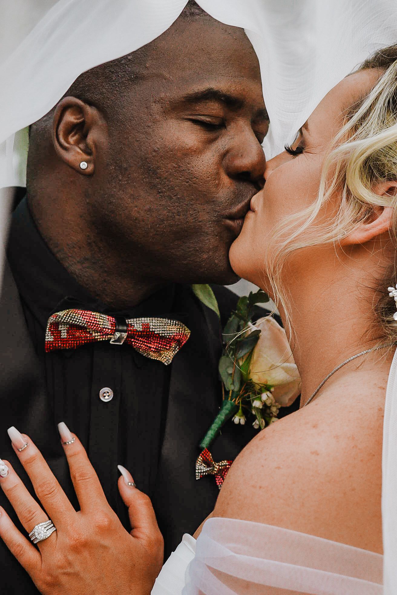 Interracial couple sharing romantic kiss under veil, groom in black suit with colorful bow tie, bride showing diamond ring.