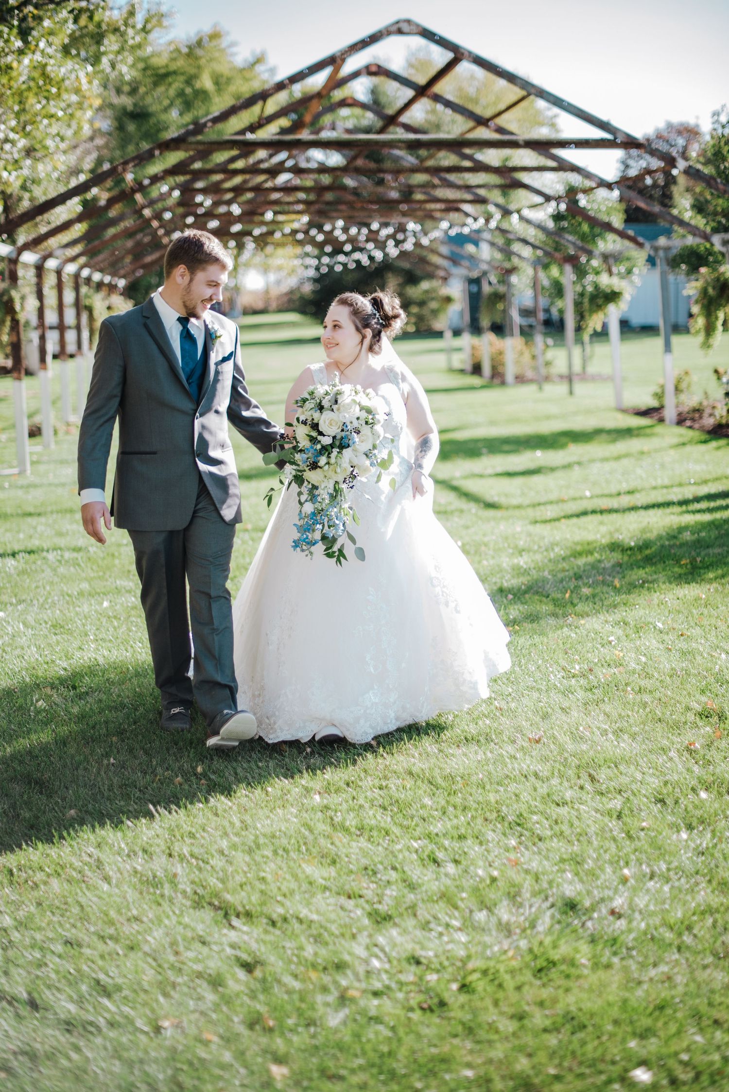 Bride and groom walk hand in hand through a garden pergola, bride holding a white floral bouquet.