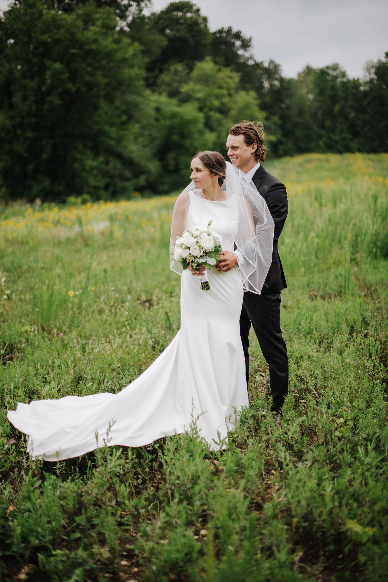 Elegant bride in minimalist white gown with cathedral train standing with groom in black suit in lush green meadow.