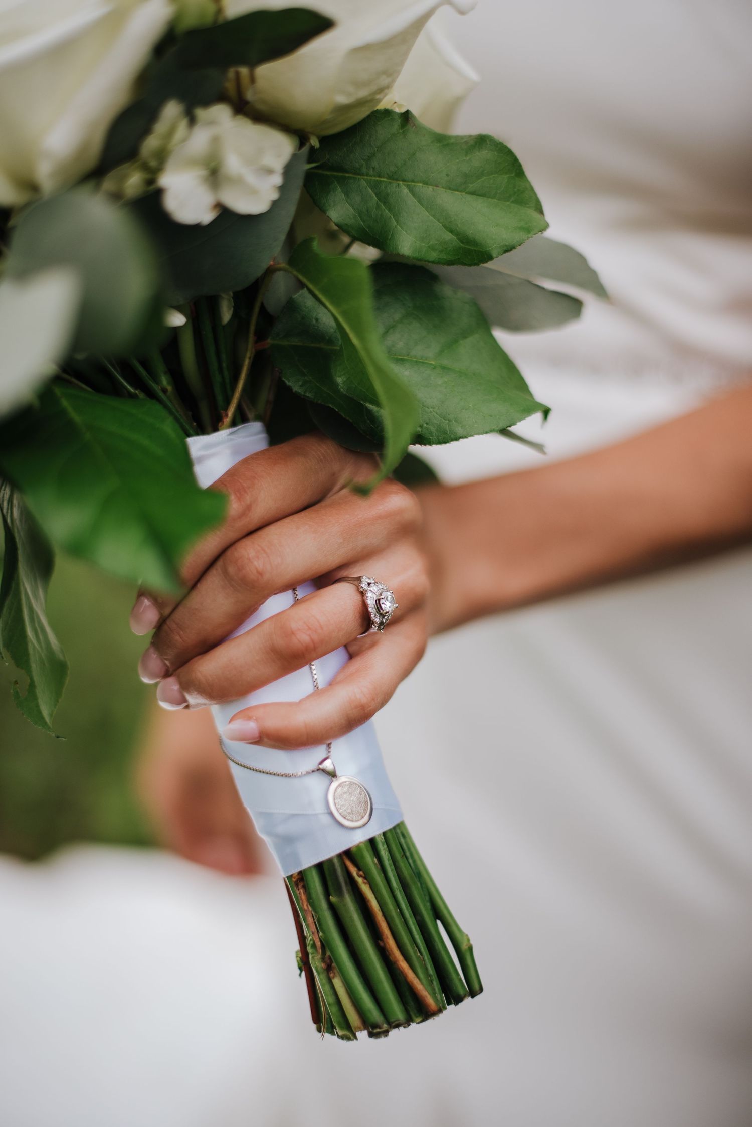 Bride holding white rose bouquet with diamond engagement ring and locket charm tied to stems.