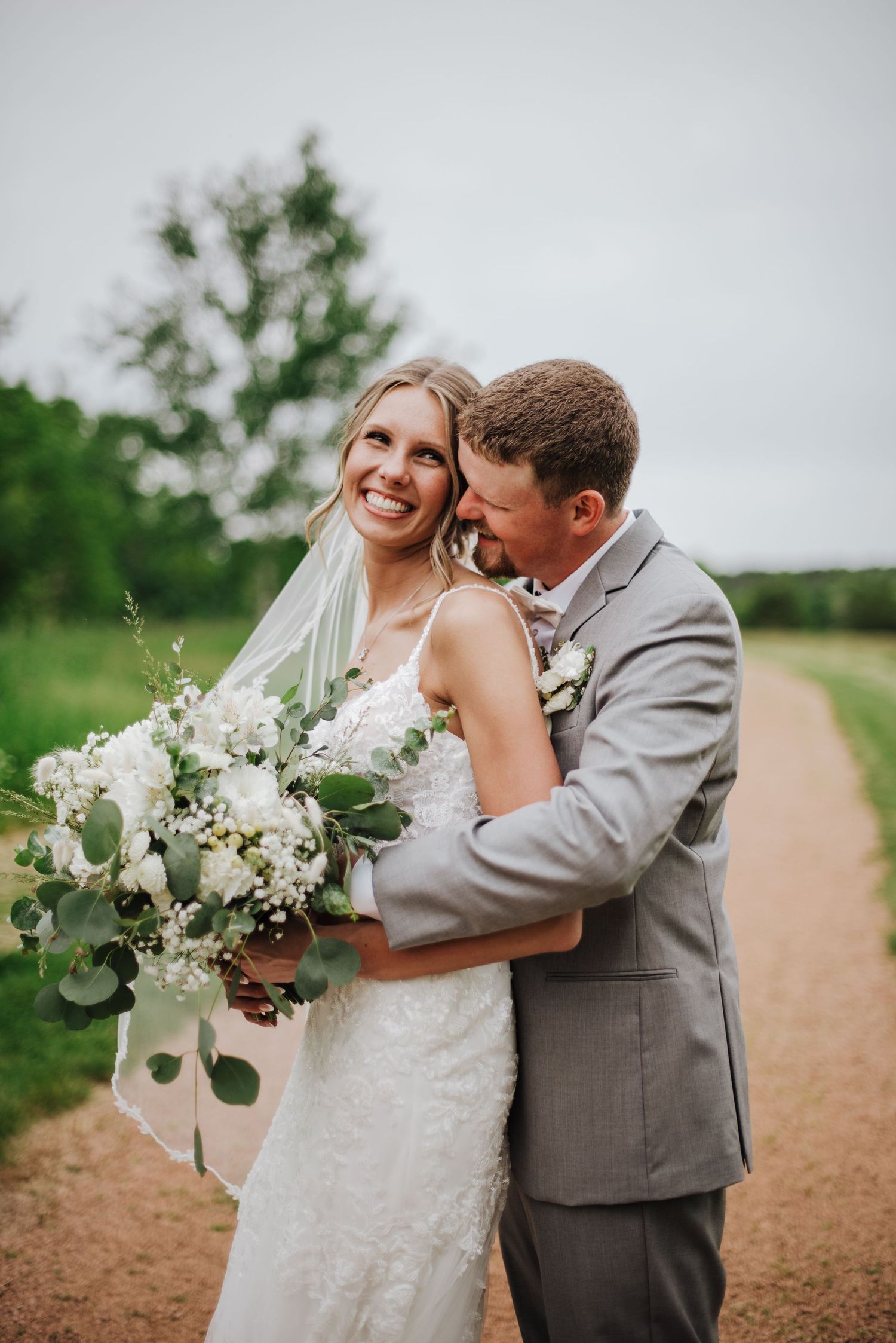 Smiling bride with white bouquet hugged by groom in gray suit on dirt path.
