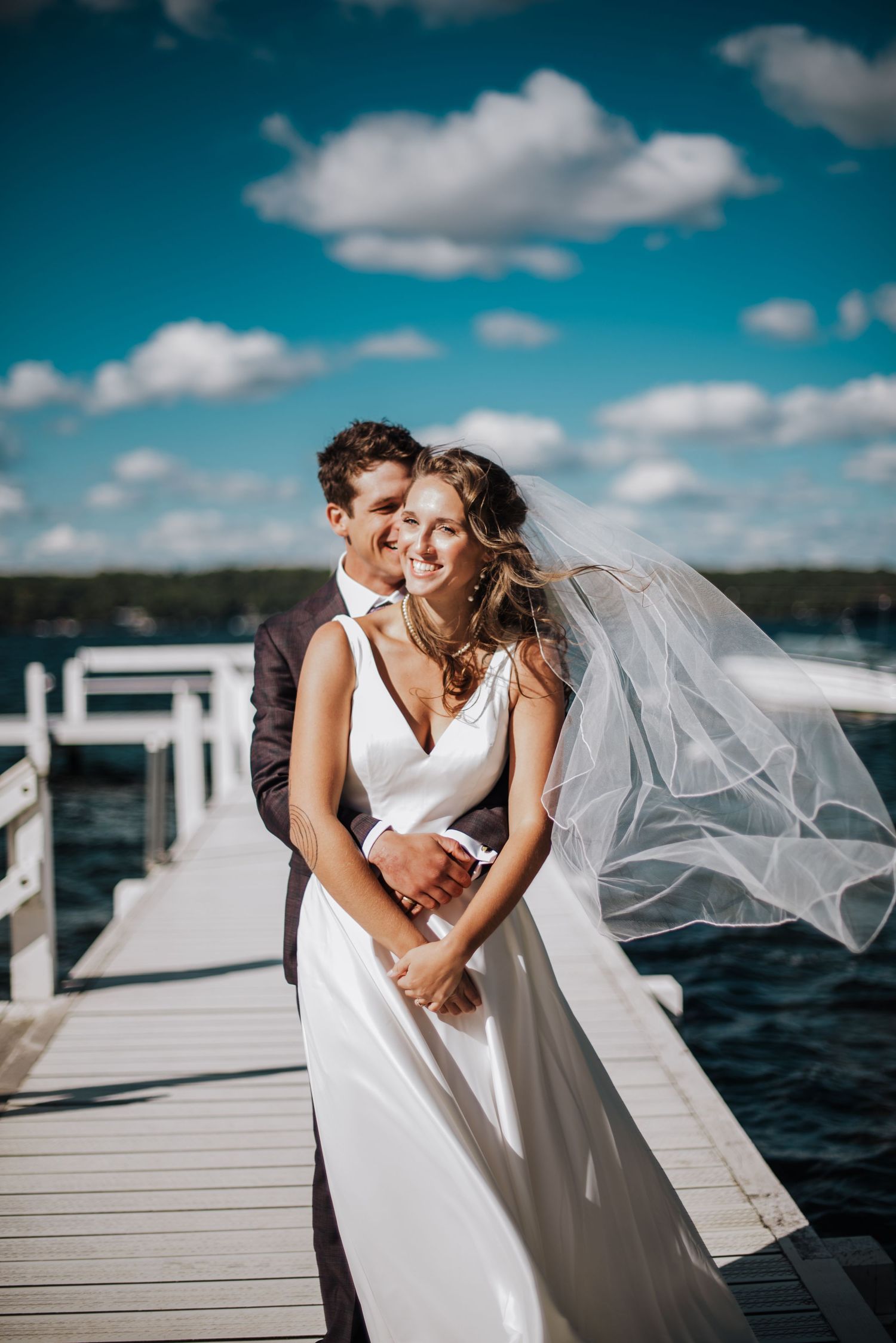 Bride and groom embrace on a lakeside dock under a blue sky, her veil flowing in the wind.