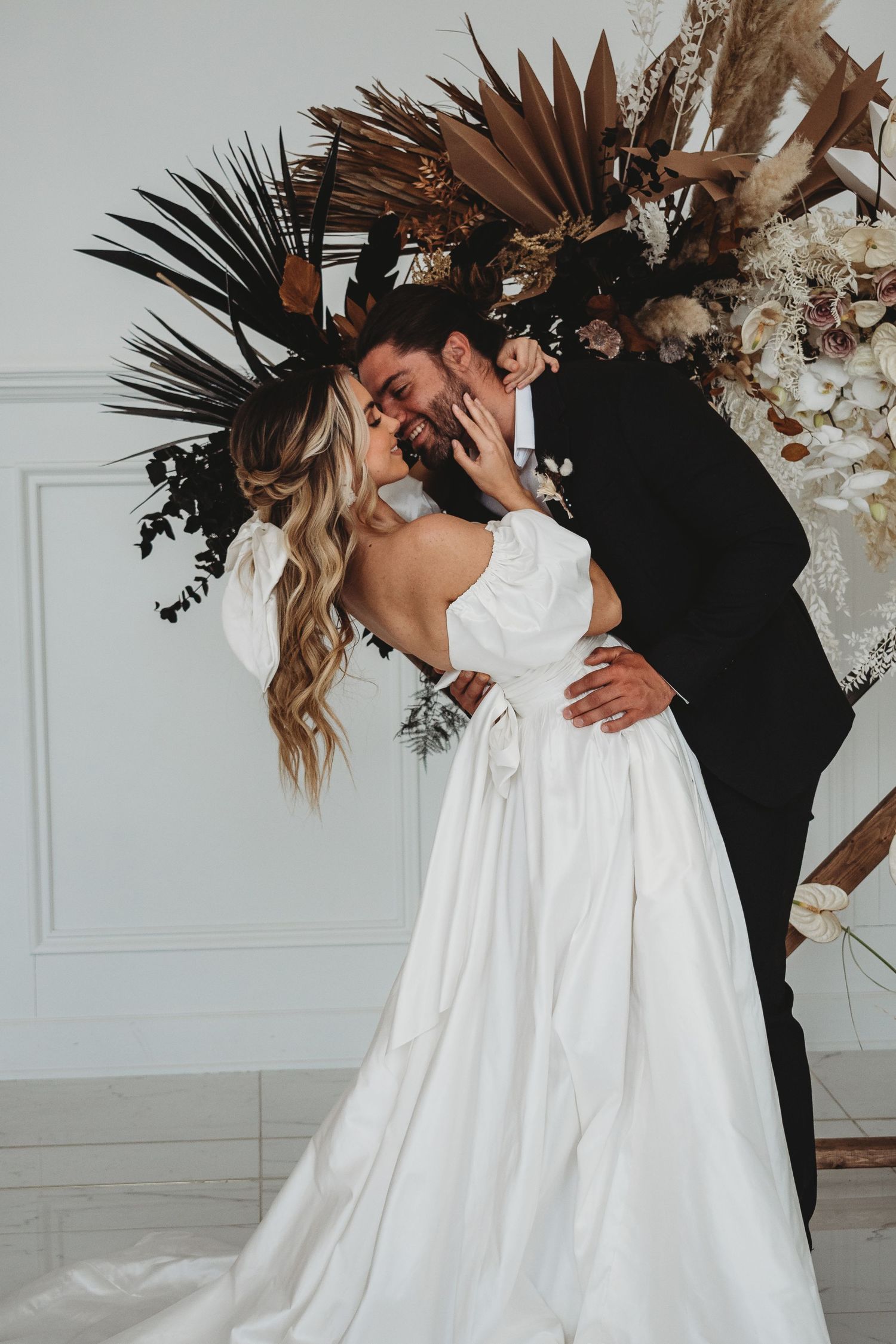 Wedding couple sharing a kiss beneath a dramatic dried botanical arch with dark palm fronds.