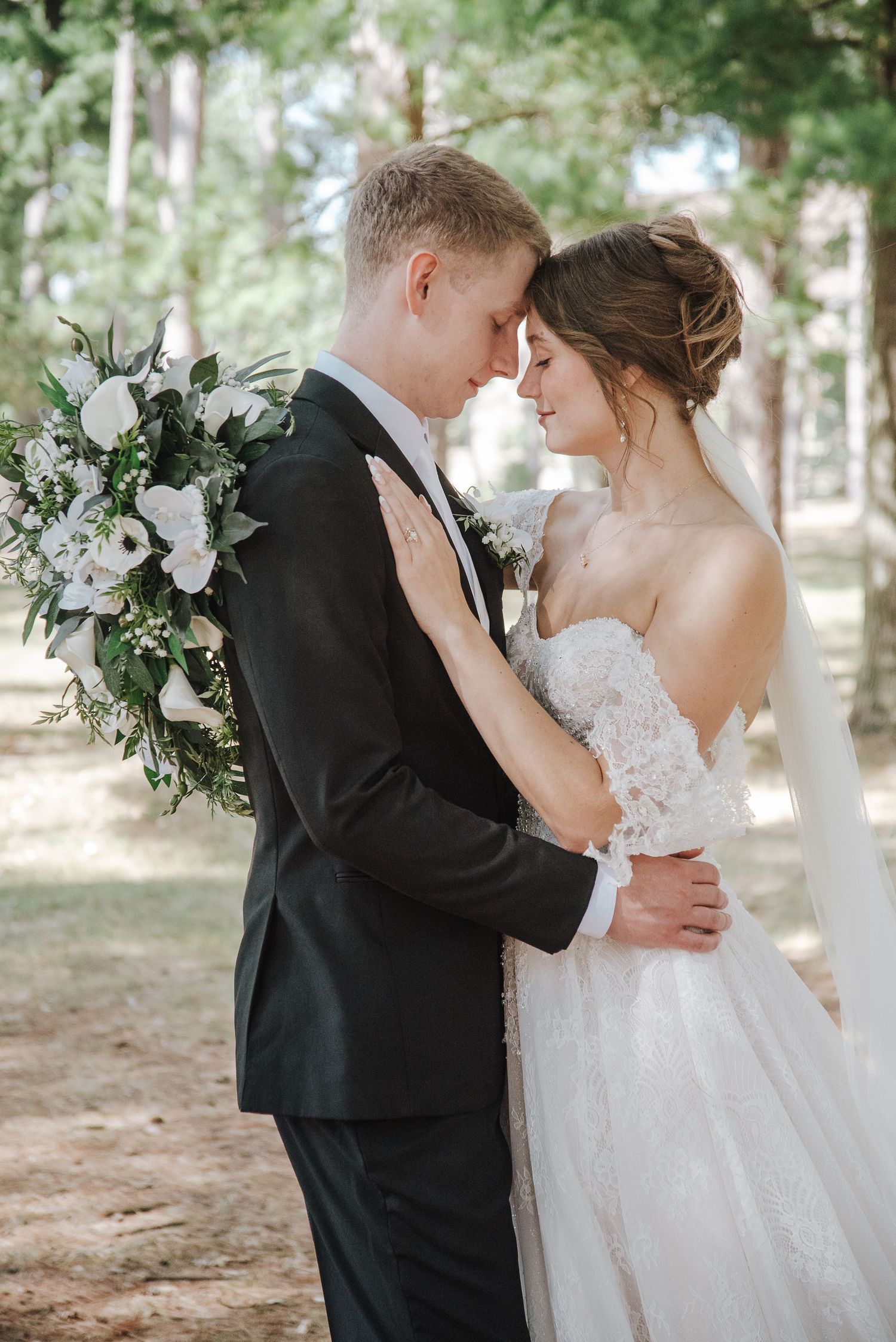 Bride and groom touching foreheads tenderly in forest setting with white floral bouquet outdoors.
