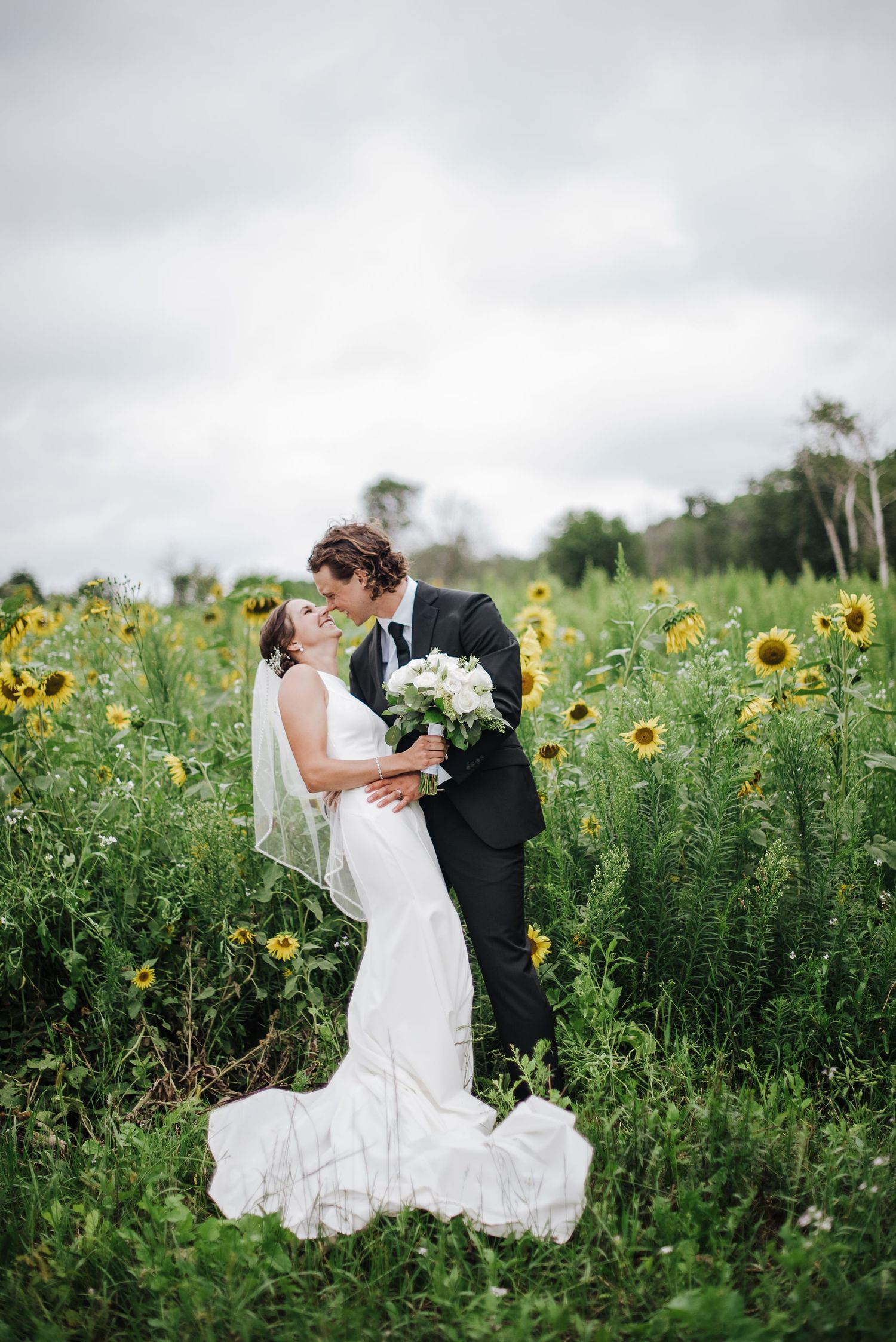 Bride and groom kissing in a sunflower field, bride in white gown with veil holding bouquet.
