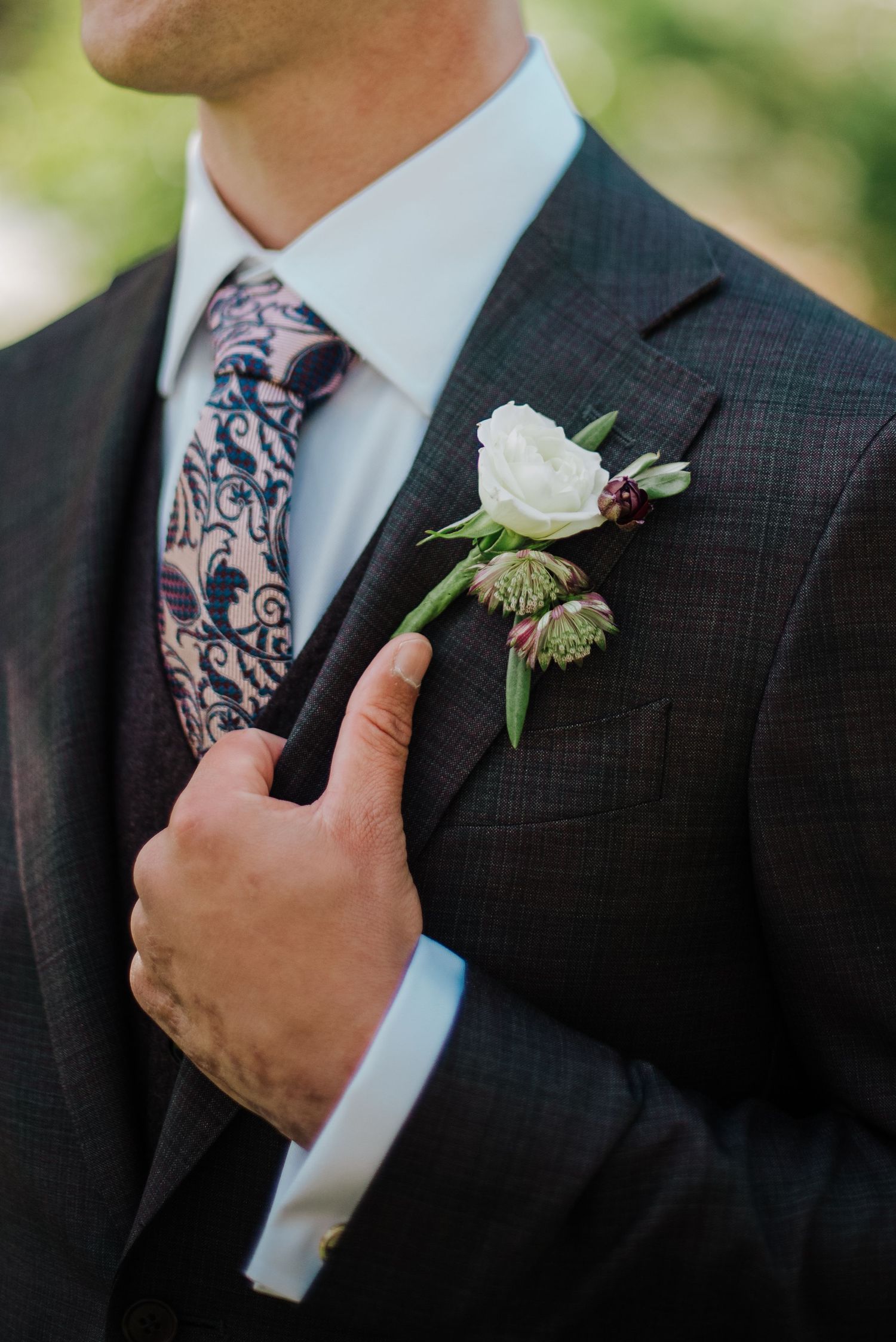 Groom in dark suit adjusting white rose boutonniere with patterned tie at wedding ceremony.