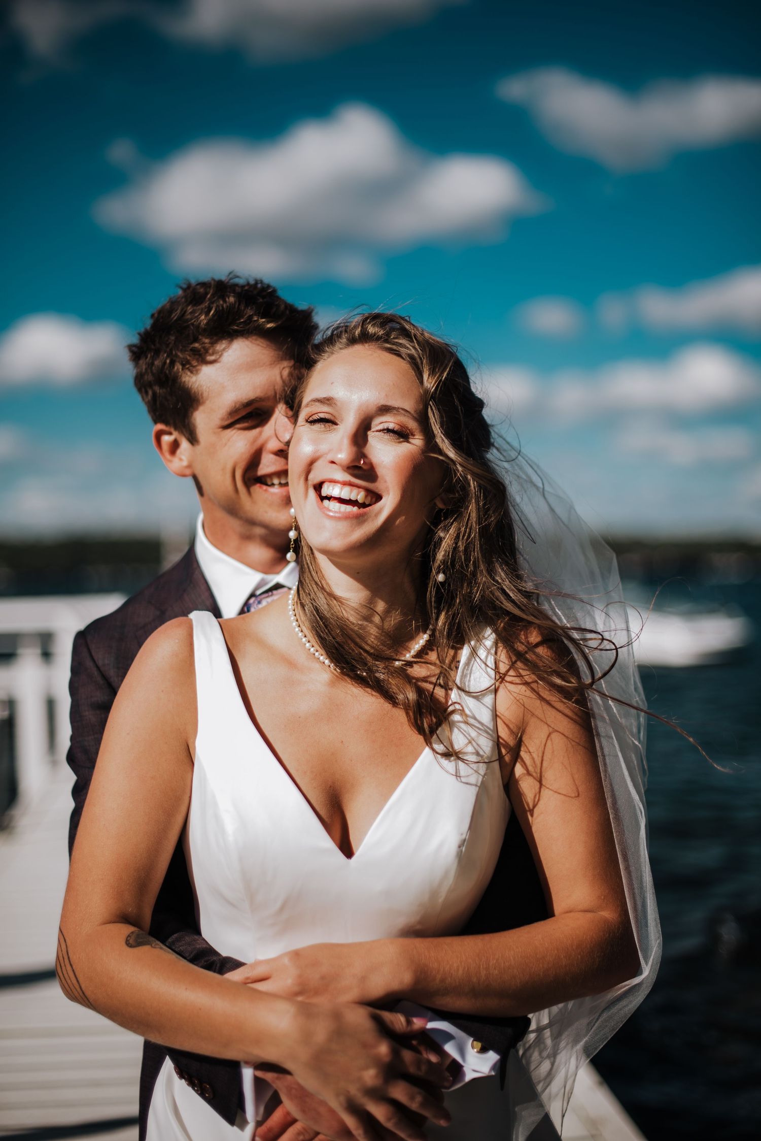 Joyful bride in white V-neck gown laughing with groom on waterfront dock under bright blue sky with veil blowing in wind.