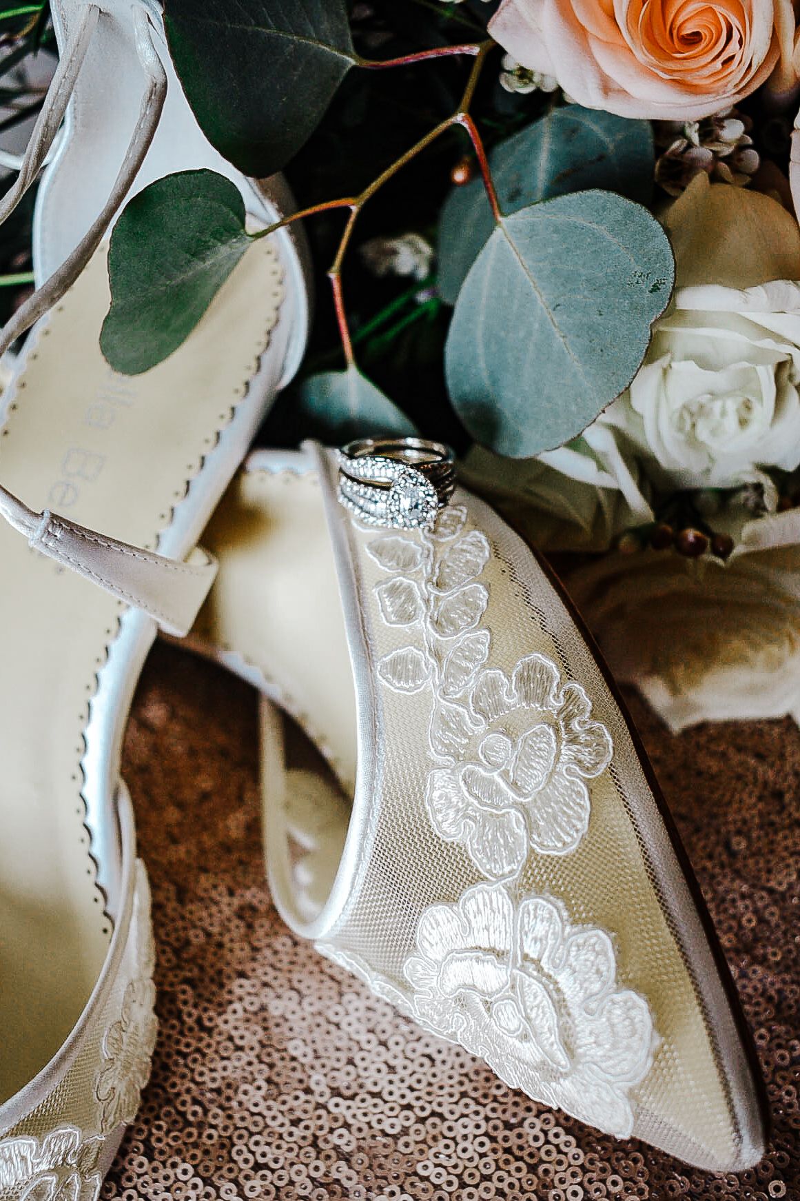 Close-up of lace bridal heels with floral embroidery and diamond wedding rings nestled near a soft floral bouquet.