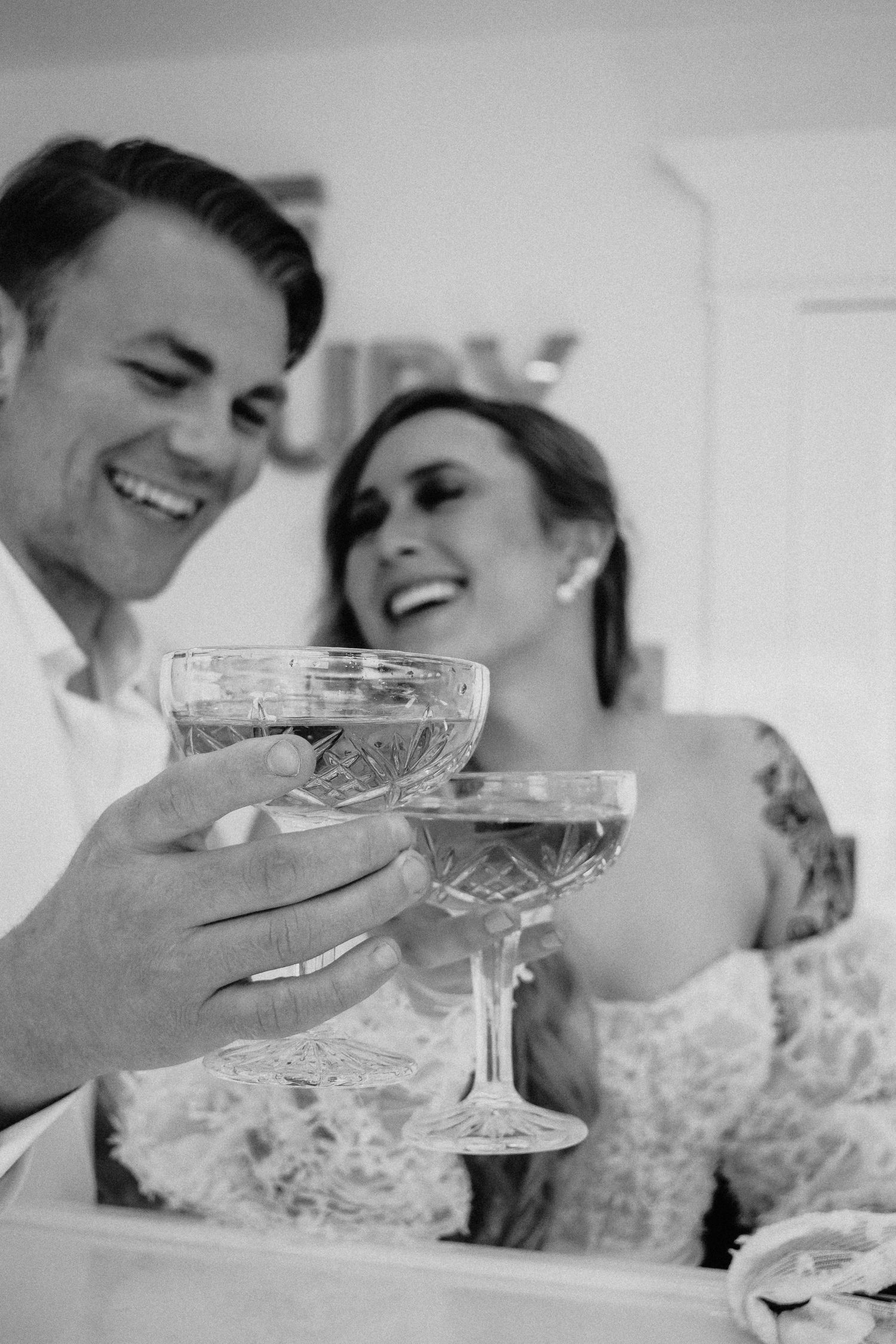 Black and white photo of laughing newlyweds toasting with crystal coupe glasses in a joyful wedding celebration.