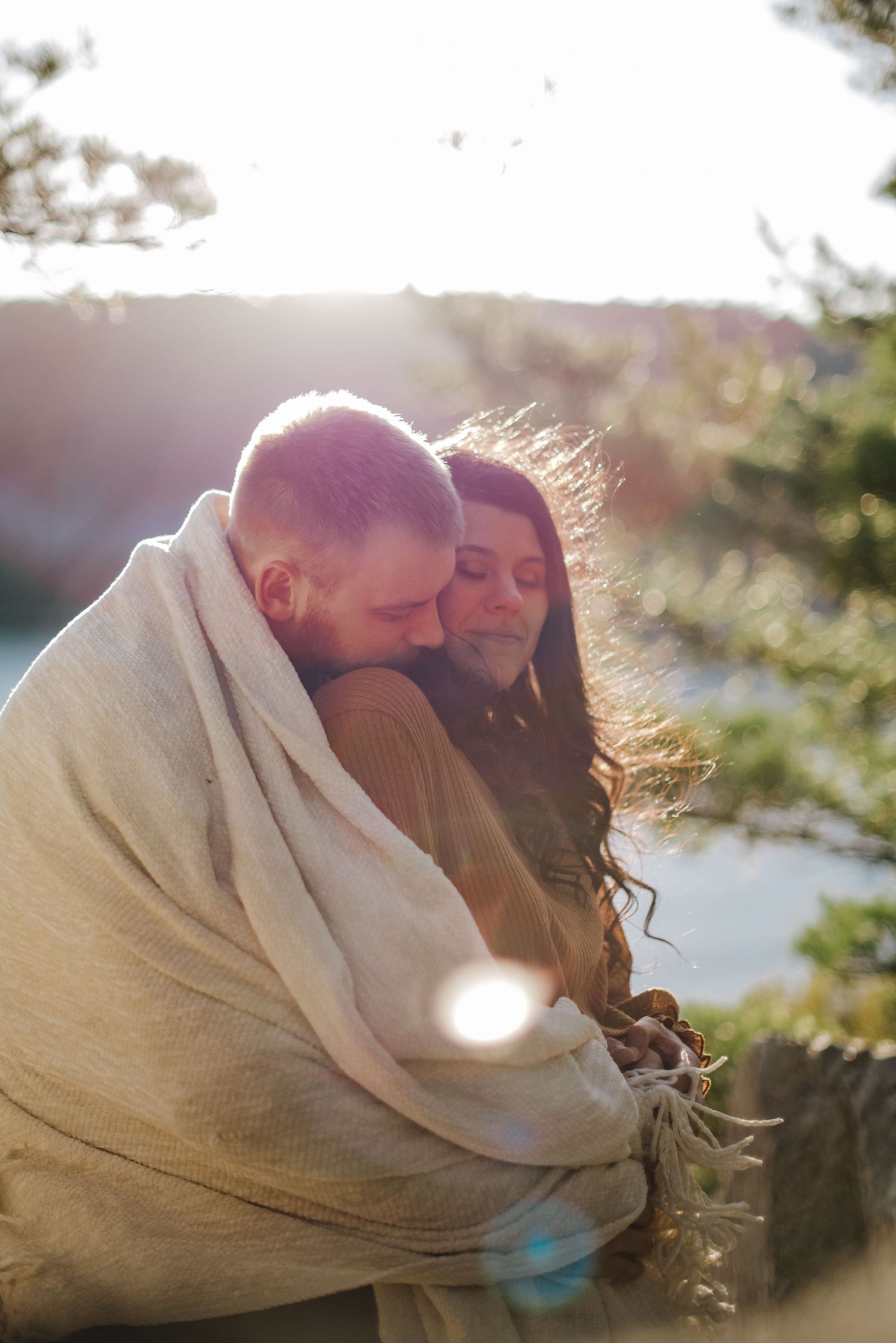 Couple wrapped in a cozy blanket together outdoors in golden sunlight near pine trees at Devil's Lake State Park in Wisconsin