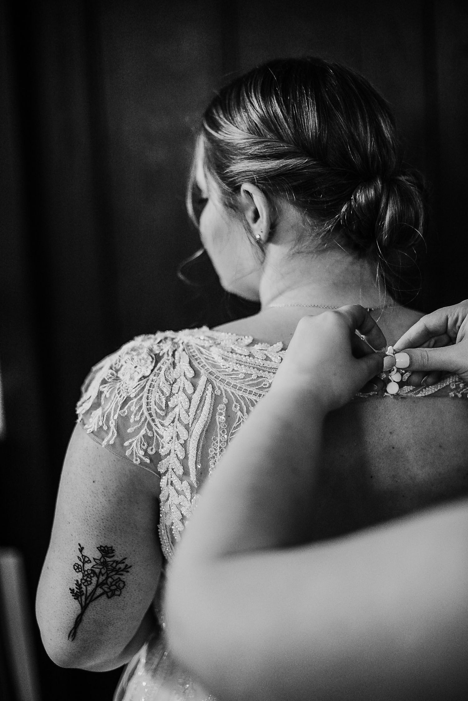Black and white photo of bride with floral lace back gown and tattoo being buttoned up.