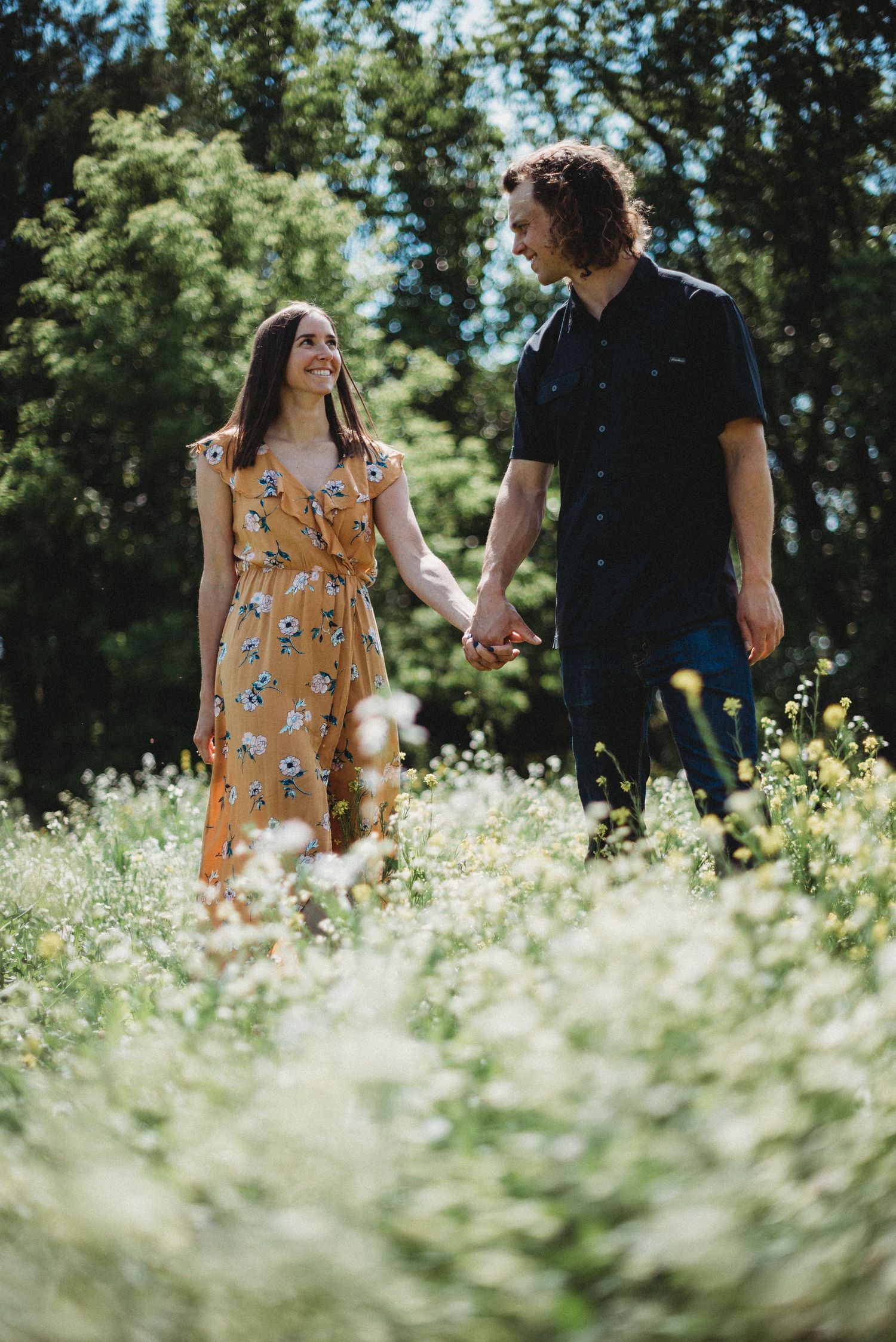 Couple holding hands and smiling at each other in a sunny wildflower meadow, woman in floral dress at home in Wisconsin