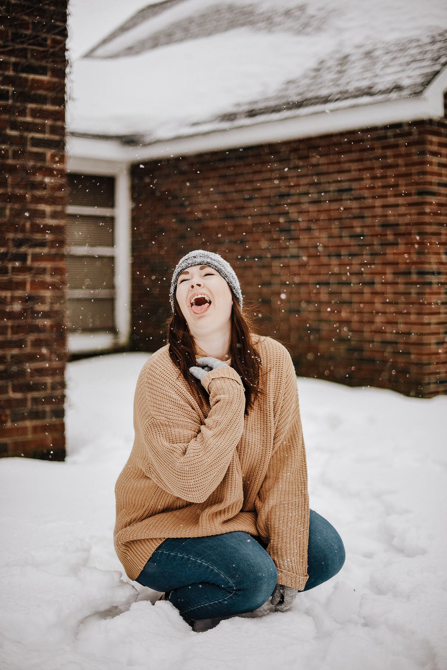 A joyful young woman in a tan sweater and beanie sits in the snow, catching snowflakes with her mouth open wide.