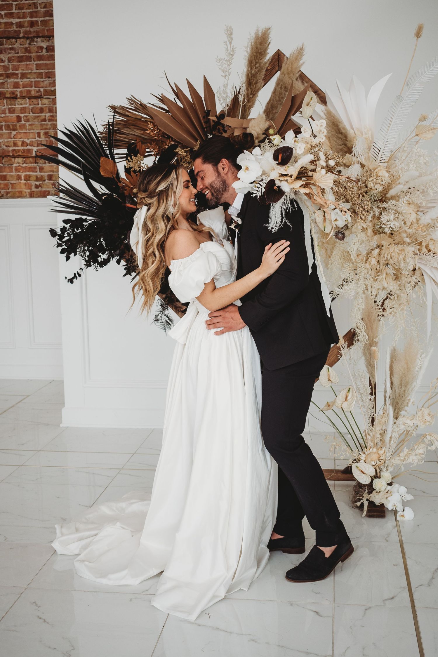 Bride and groom share a romantic moment before a boho ceremony arch adorned with dried palms and pampas grass.