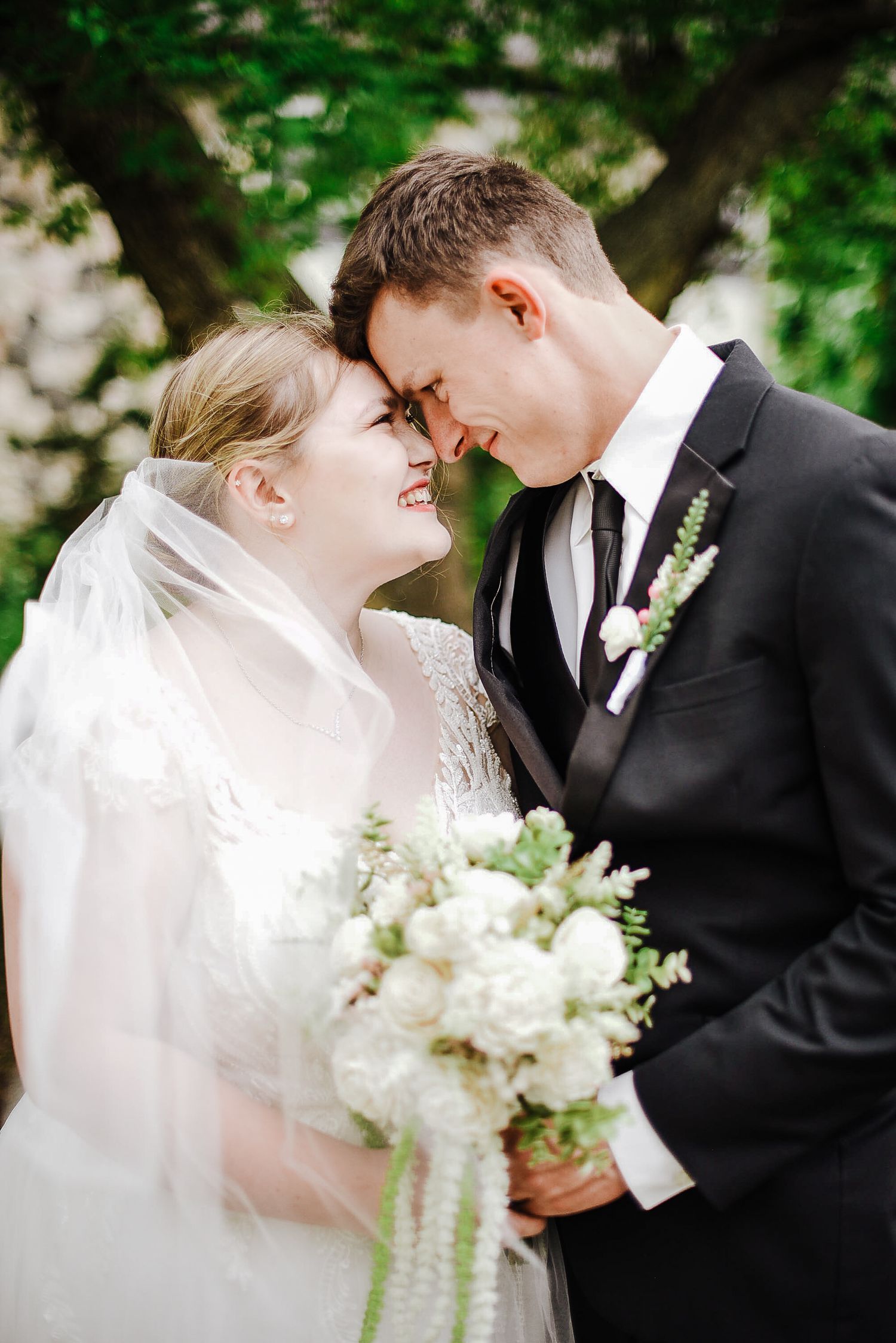 Bride and groom touching foreheads tenderly under sheer veil, bride holding white bouquet with green ribbons outdoors.