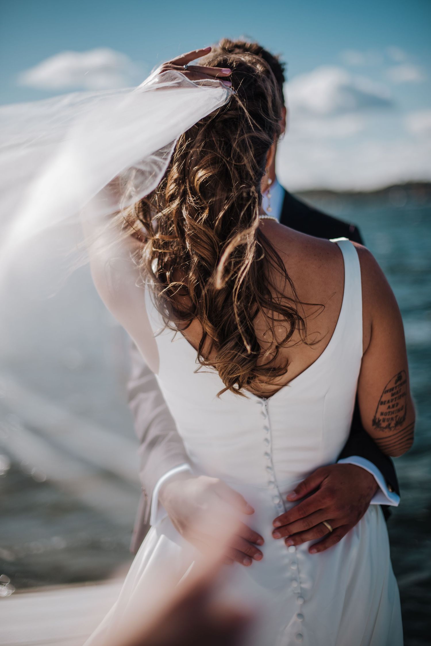 Romantic back view of bride in white button-back gown with flowing veil blowing in wind near waterfront, groom embracing her.