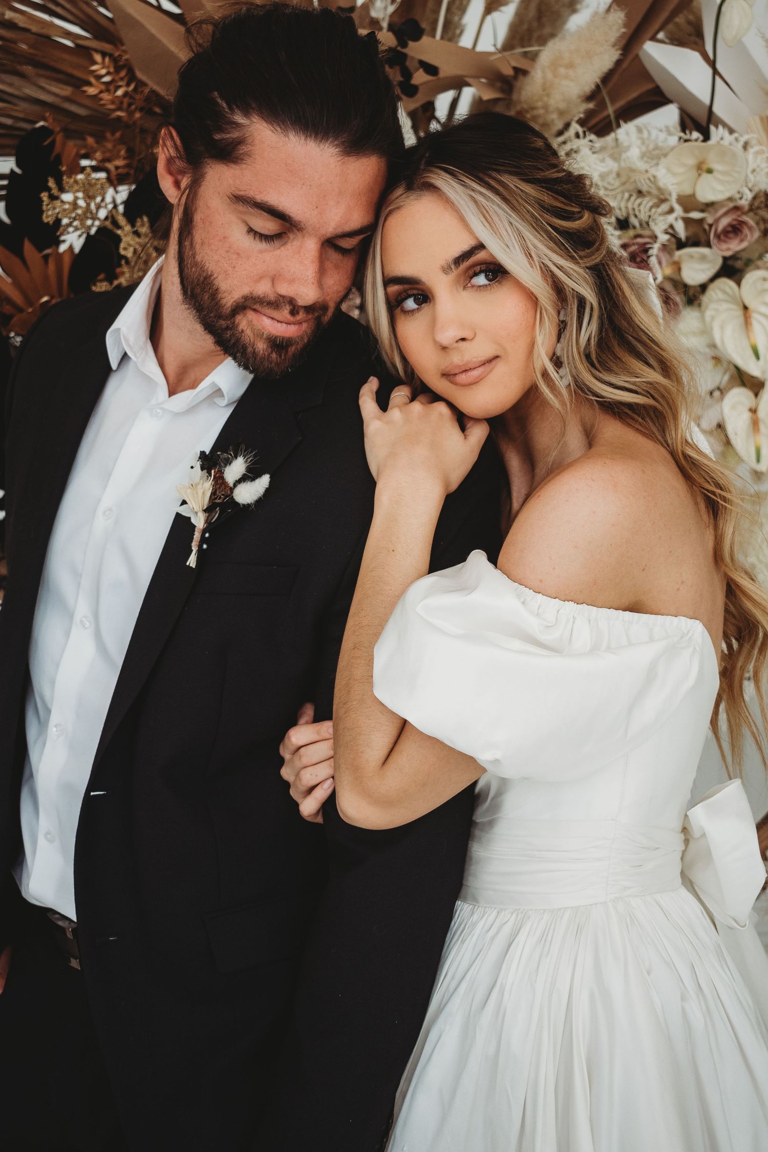 Elegant bride in off-shoulder gown and groom in black suit posing before a boho floral backdrop.