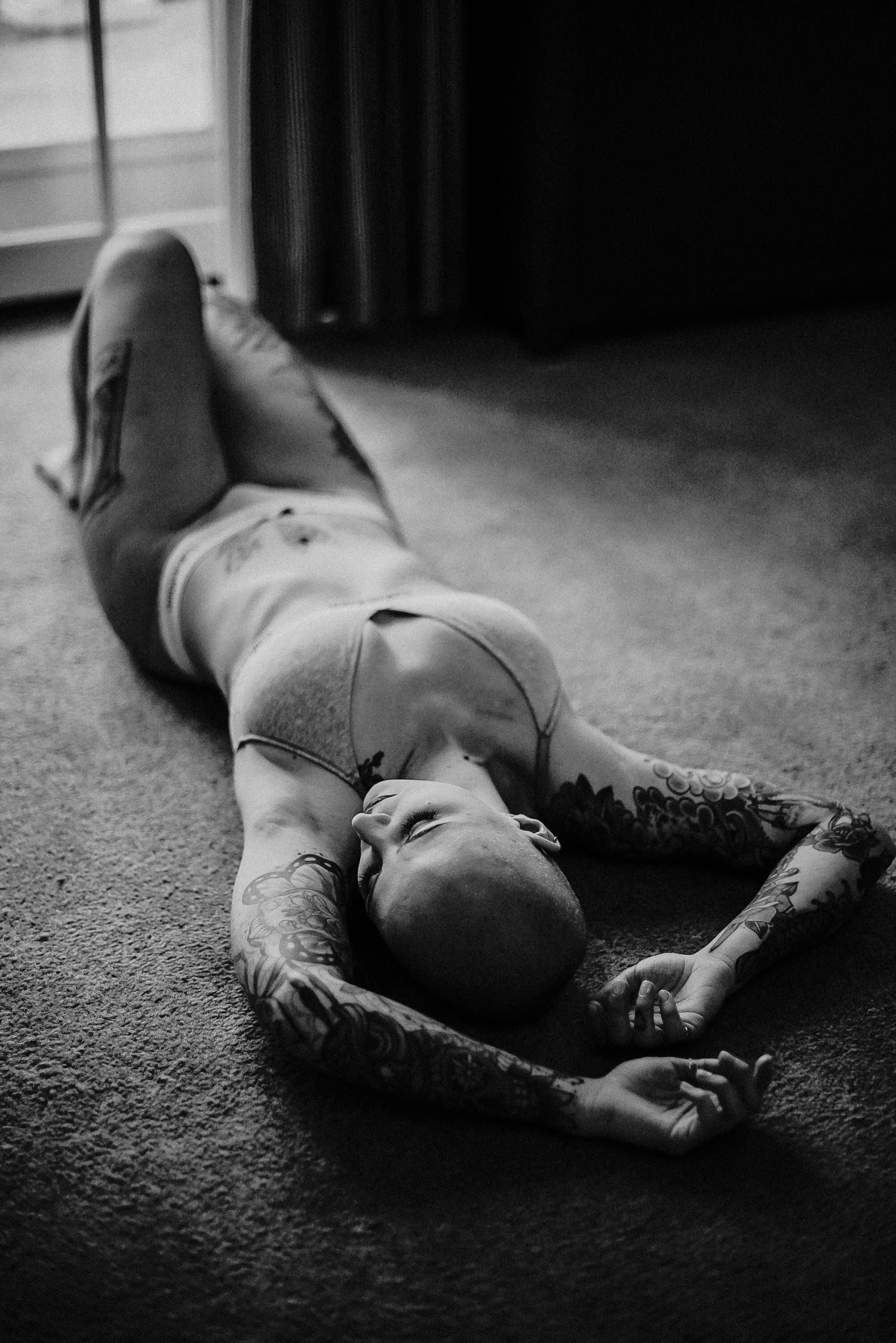 Black and white photo of a tattooed person with a shaved head lying on a carpeted floor beside a window, arms stretched overhead.