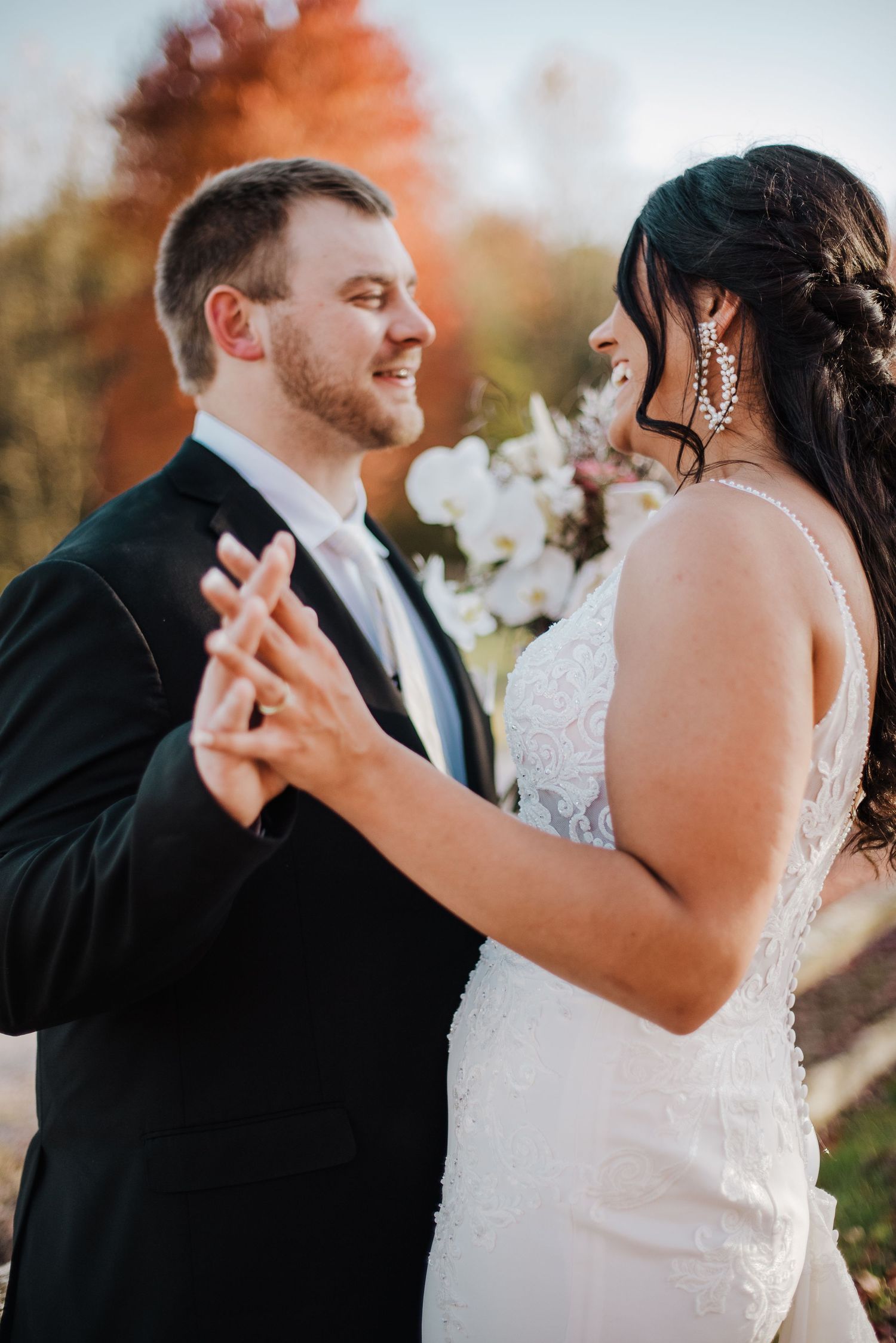 Smiling groom in black tuxedo dancing with bride in beaded gown wearing crystal earrings in golden autumn sunlight.