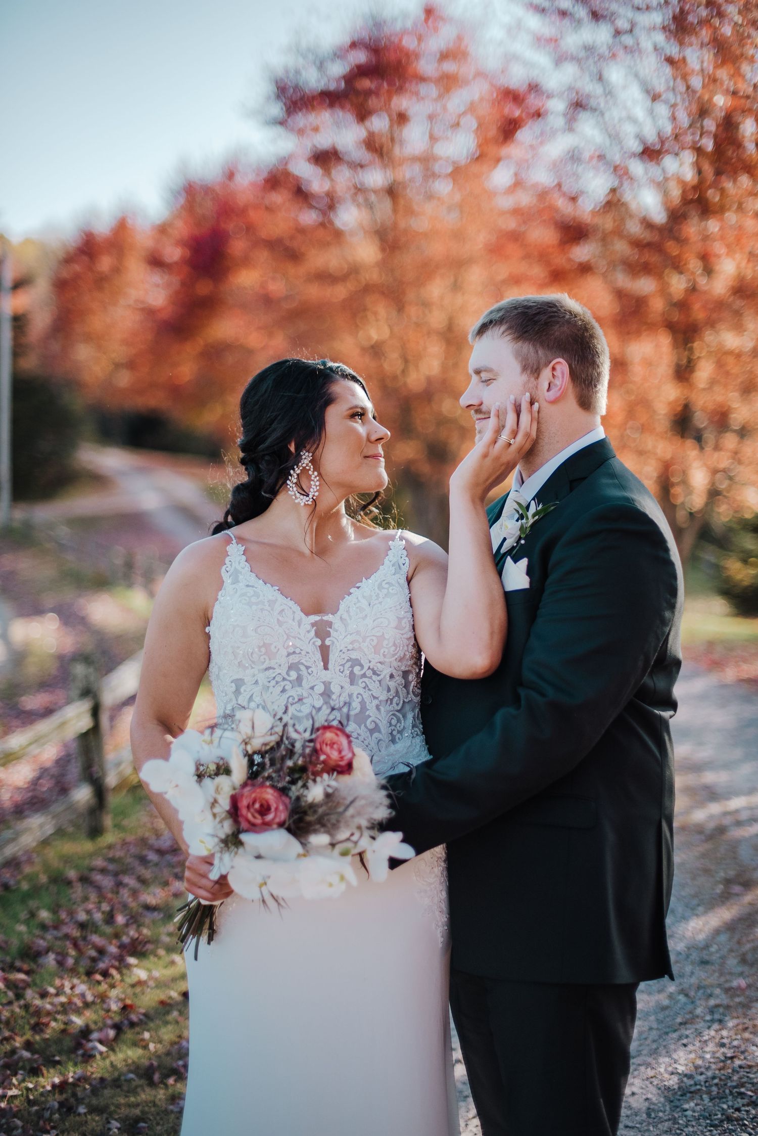 Bride tenderly cups grooms face amidst vibrant autumn foliage, holding a pink and white bouquet.