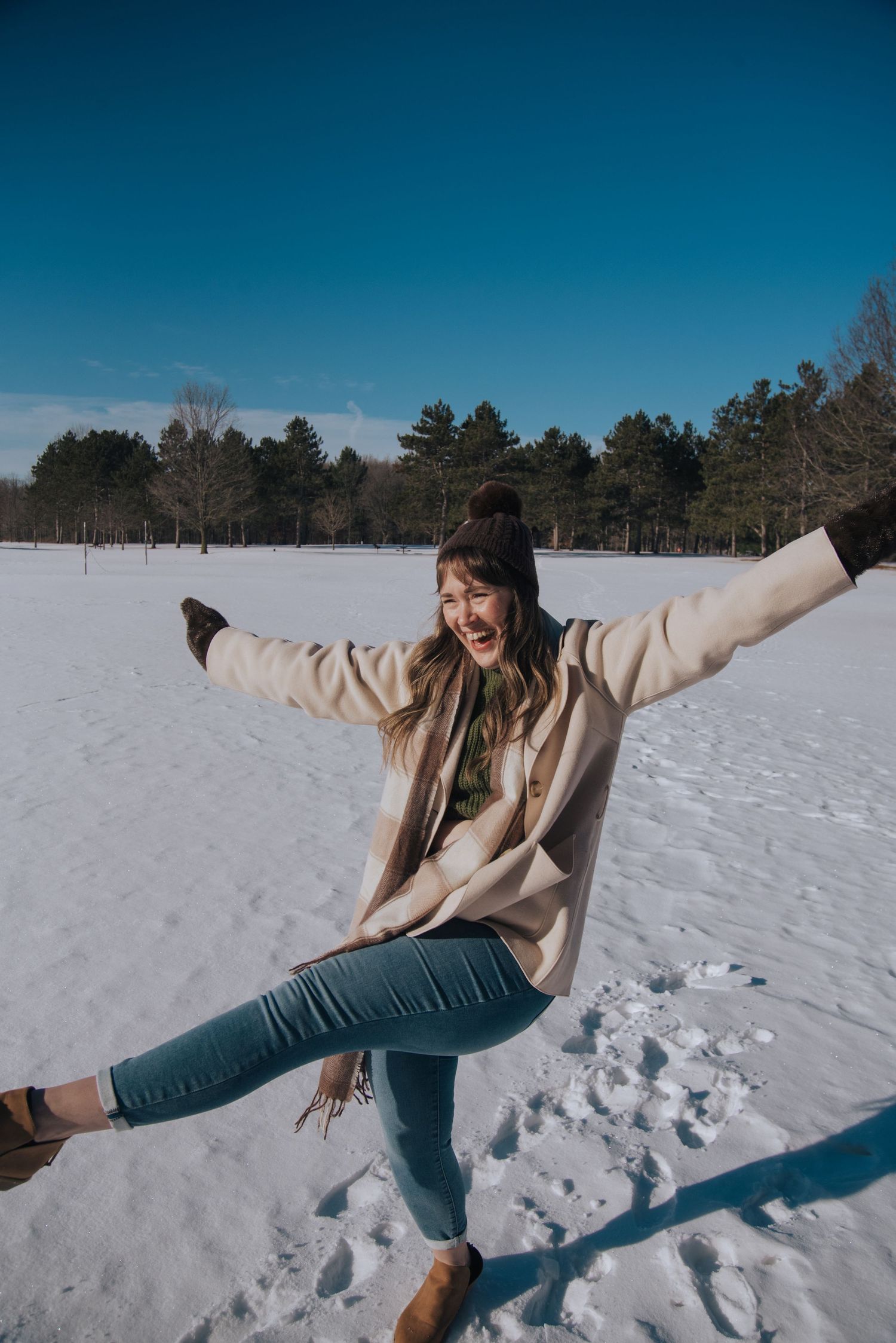 Woman in beige coat and jeans joyfully posing with arms out in a snowy winter landscape on a sunny day.