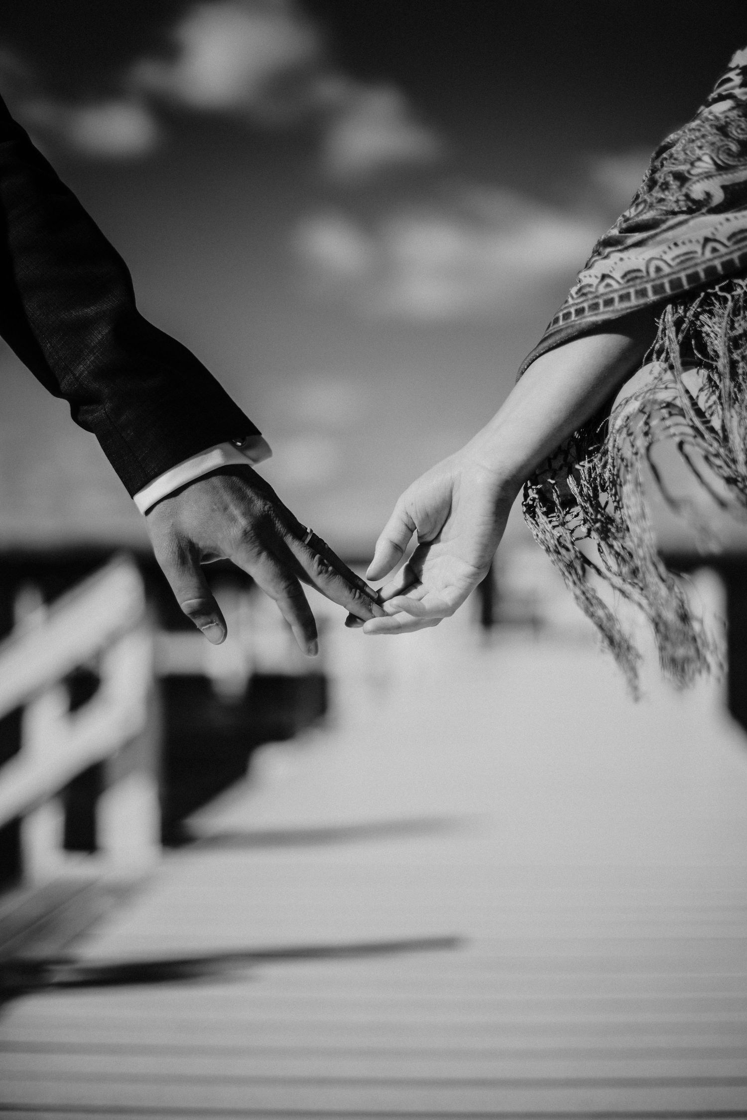 Black and white photo of couple gently touching fingertips on wooden boardwalk under blue sky in Fontana, Wisconsin