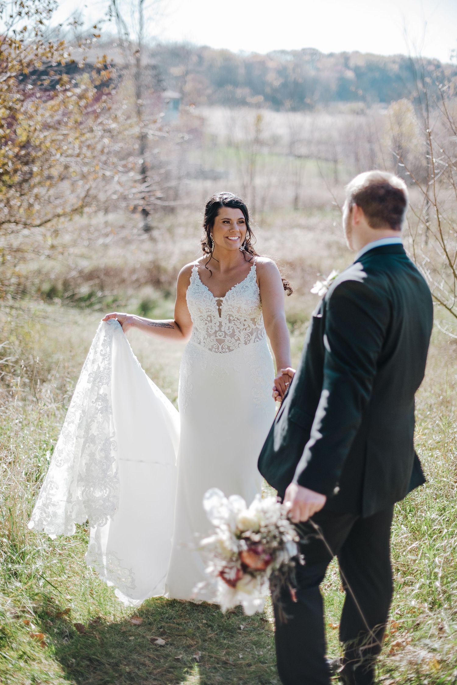 Bride in lace V-neck wedding gown smiling at groom holding bouquet in golden autumn meadow landscape setting.