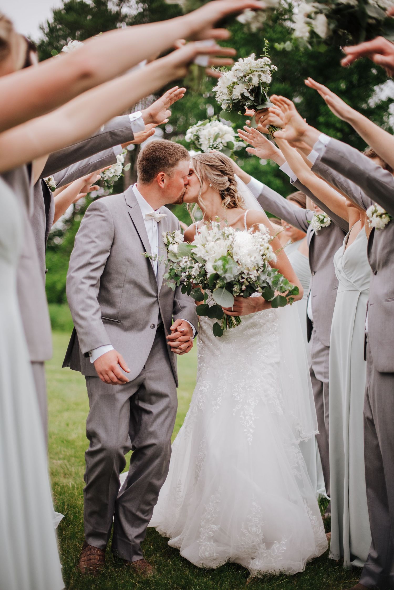 Wedding party tunnel kiss with bride in lace gown and groom in gray suit, surrounded by raised hands and white floral bouquets.