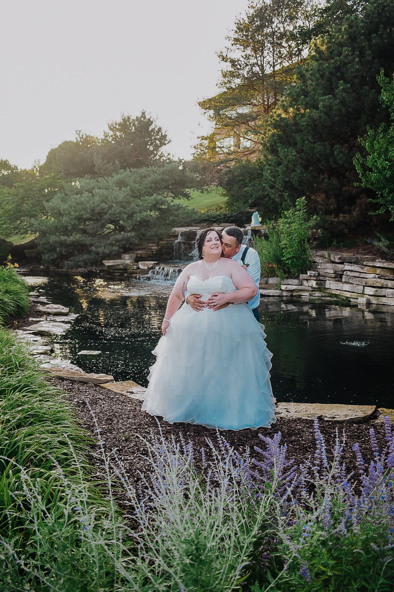 Bride and groom embrace by a serene garden pond with lavender blooms and waterfall backdrop at golden hour.