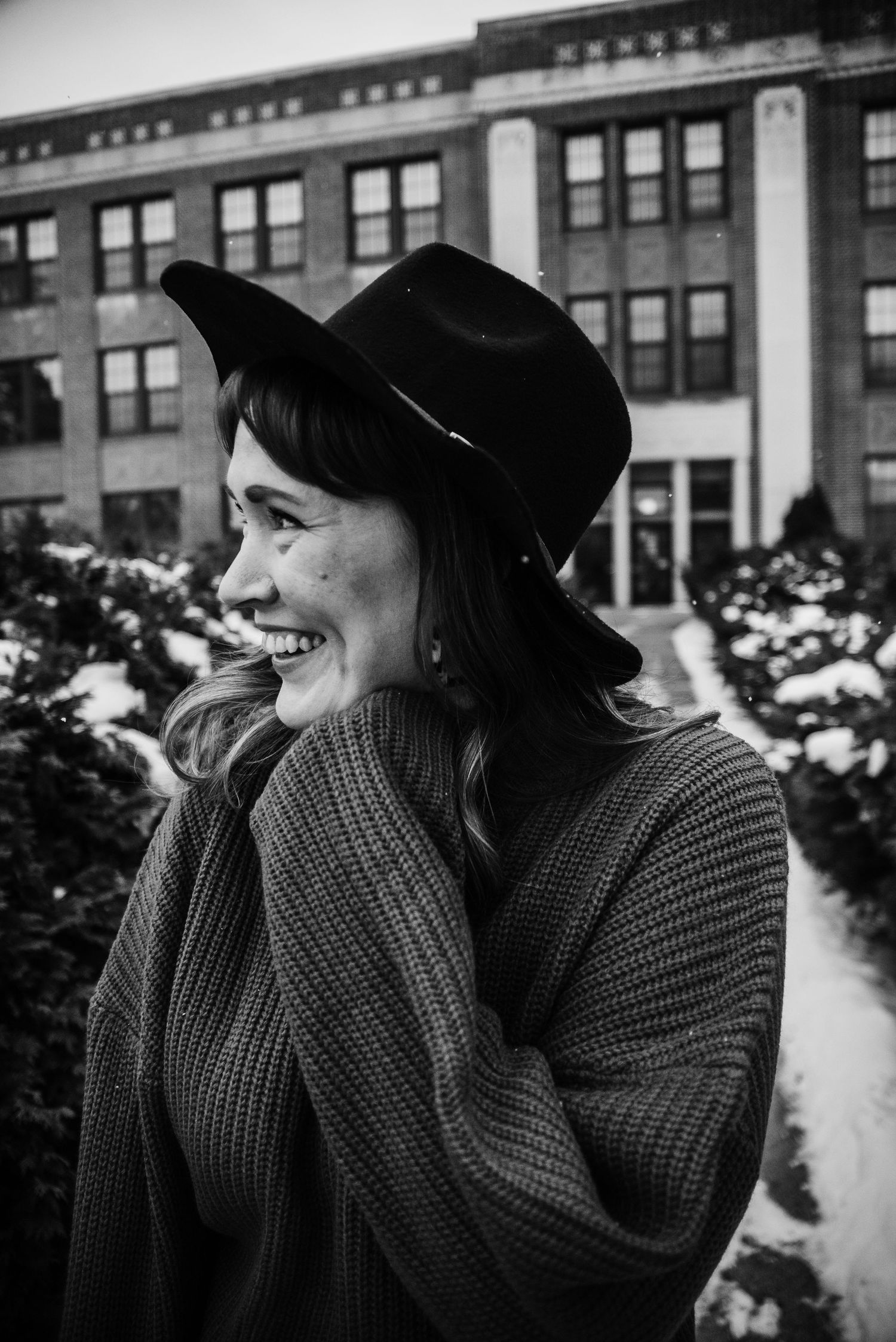 Black and white portrait of laughing woman in wide-brim hat and knit wrap outside brick building in snow.