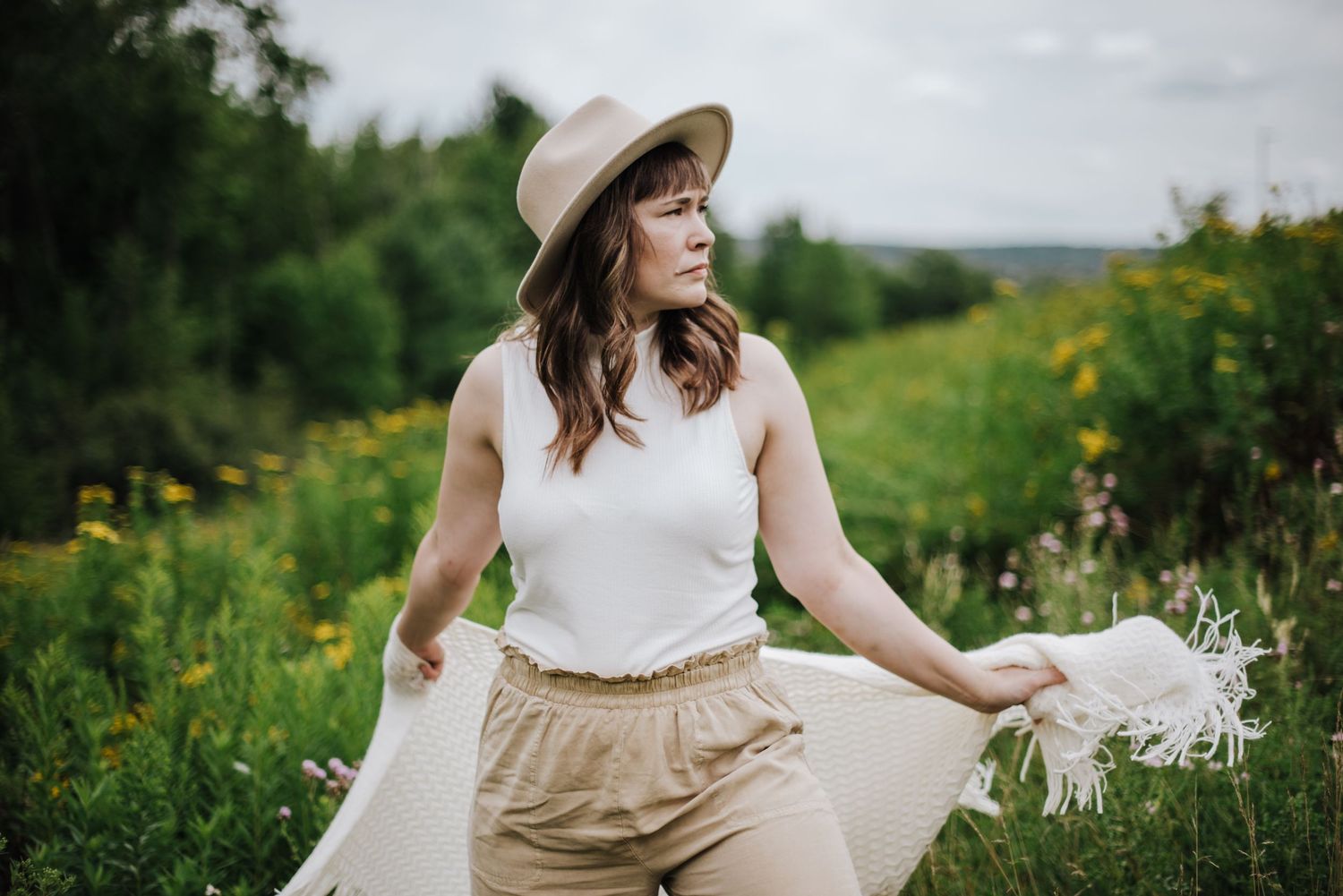 Woman in white top, beige pants, and hat holding a scarf in a lush green wildflower meadow.