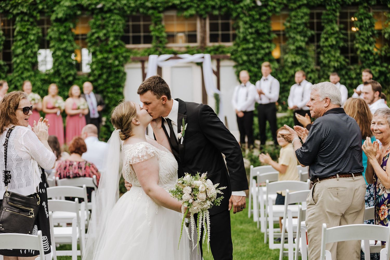 Newlyweds share their first kiss as bride and groom walk down the aisle at an outdoor garden wedding ceremony.