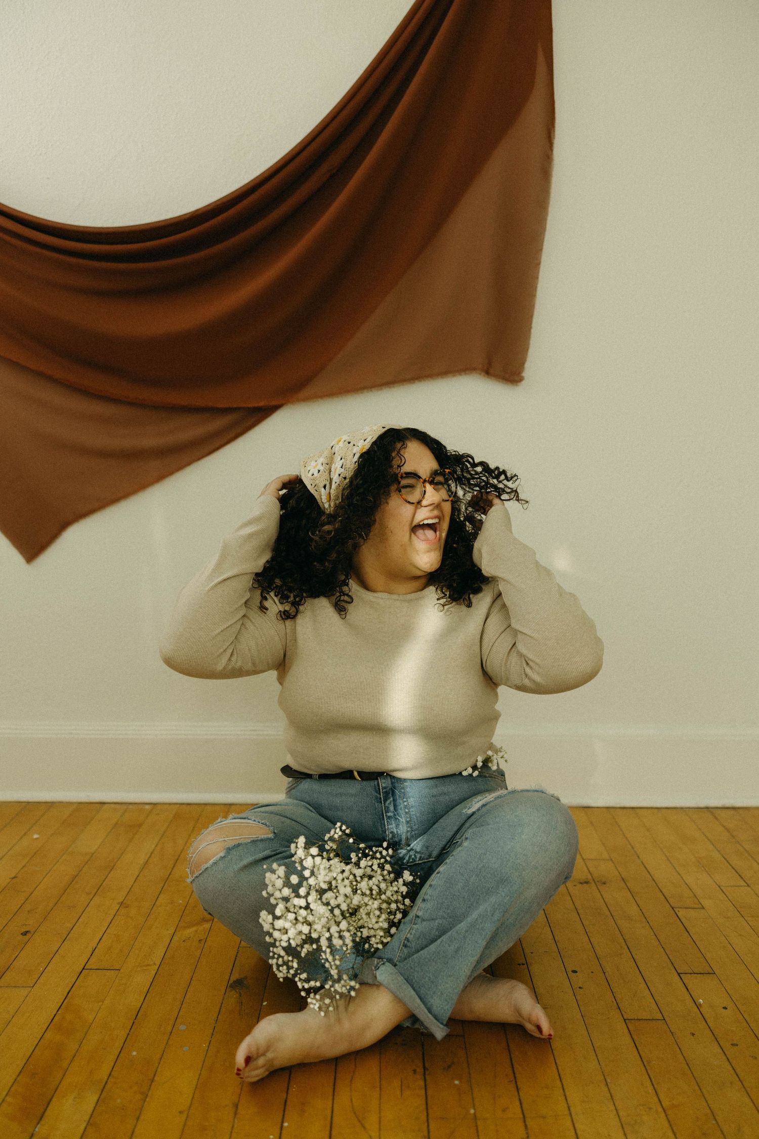 A person in a beige sweater and jeans sits cross-legged on a wooden floor with white baby's breath flowers.