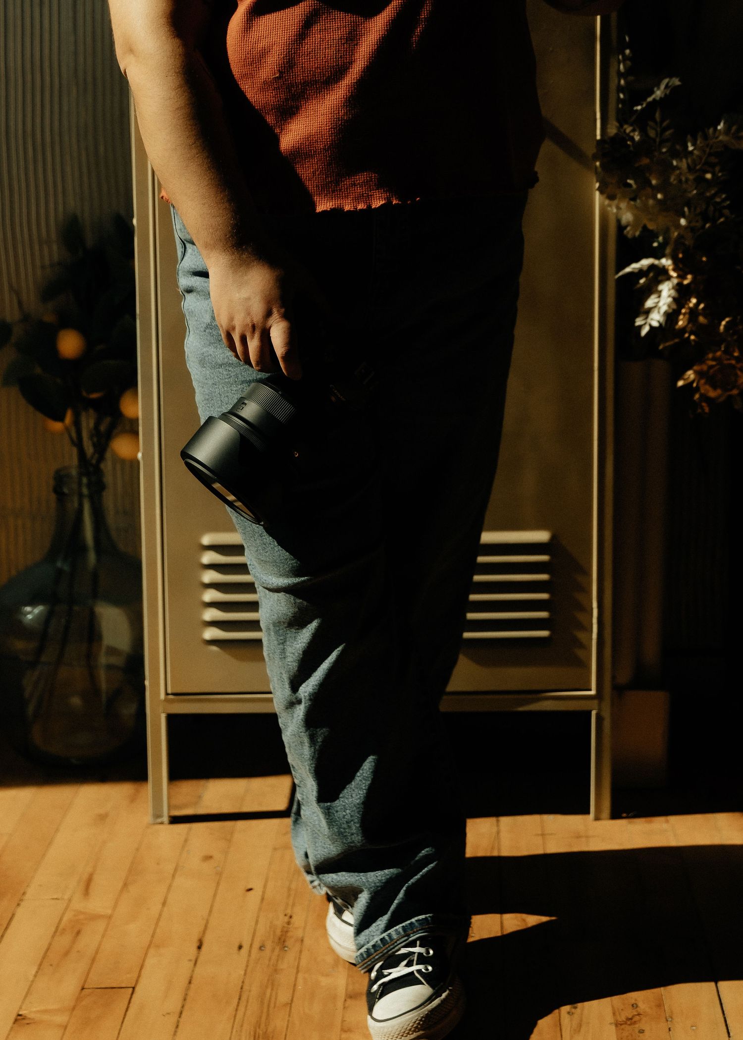 A person wearing jeans and black sneakers stands on a wooden floor with dramatic shadows cast by sunlight.