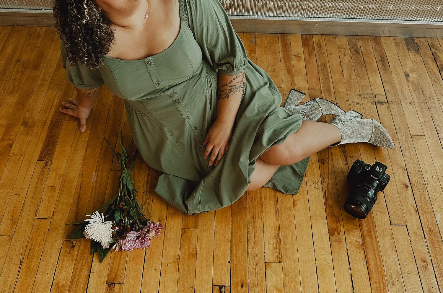 Person in olive green dress sits on hardwood floor in a relaxed pose with a camera and flowers.