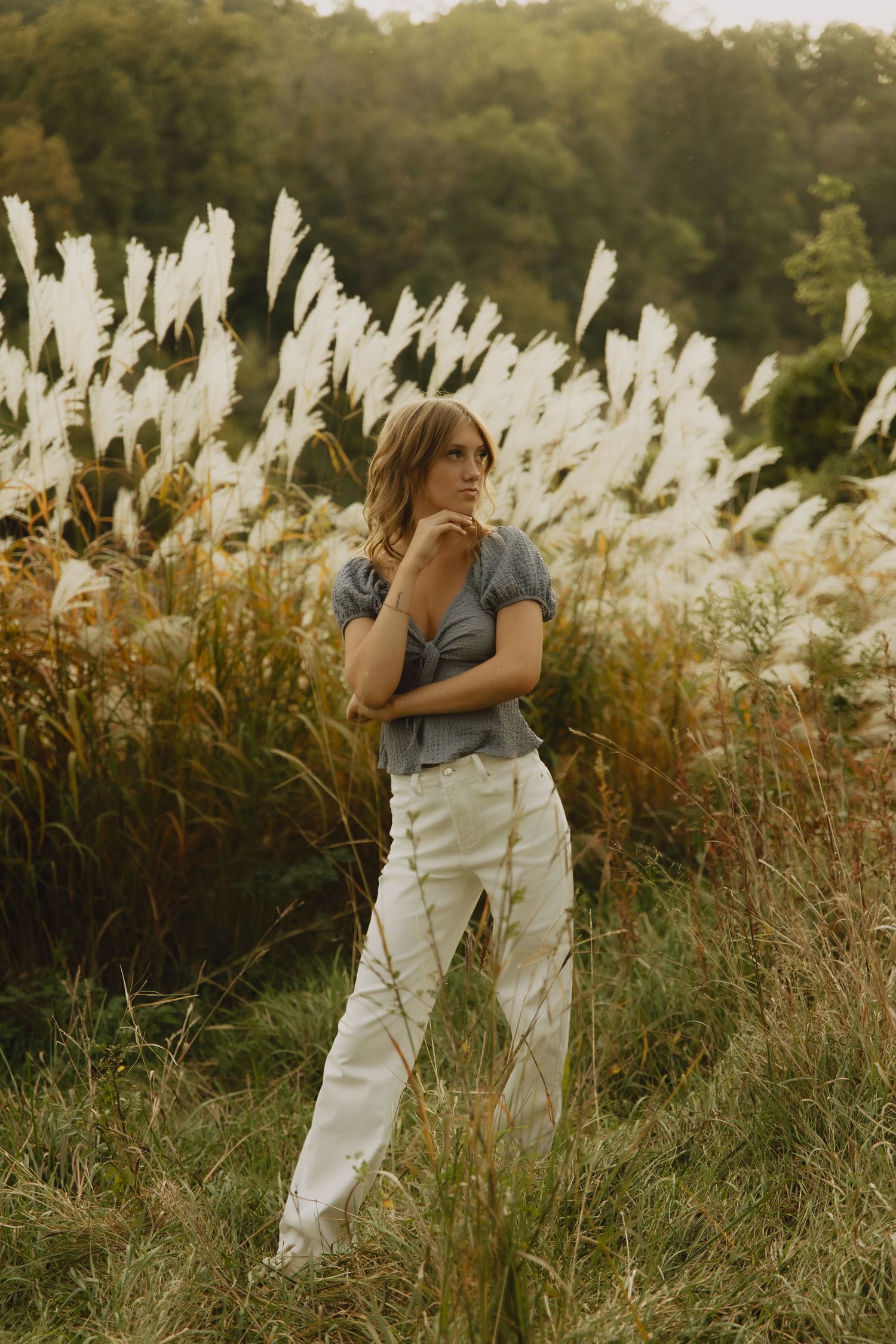 Person in white overalls and blue shirt poses thoughtfully among tall pampas grass in golden afternoon light.