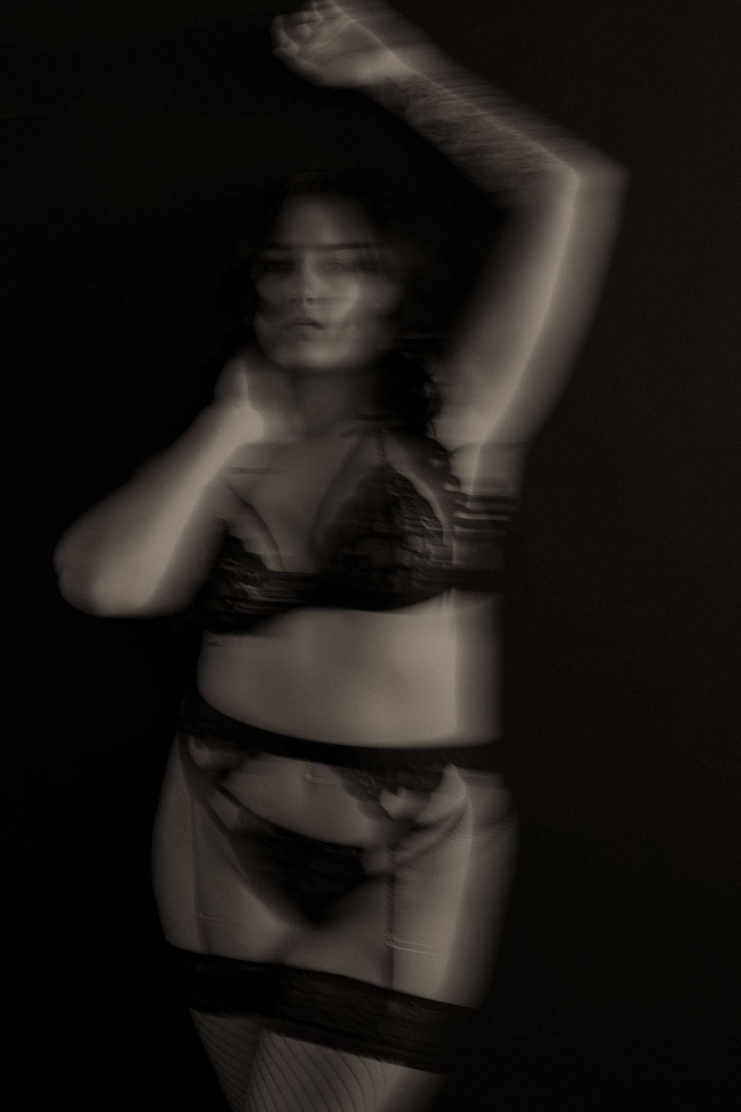 Black and white artistic blur photo of a woman in lace lingerie posing gracefully against a dark background.