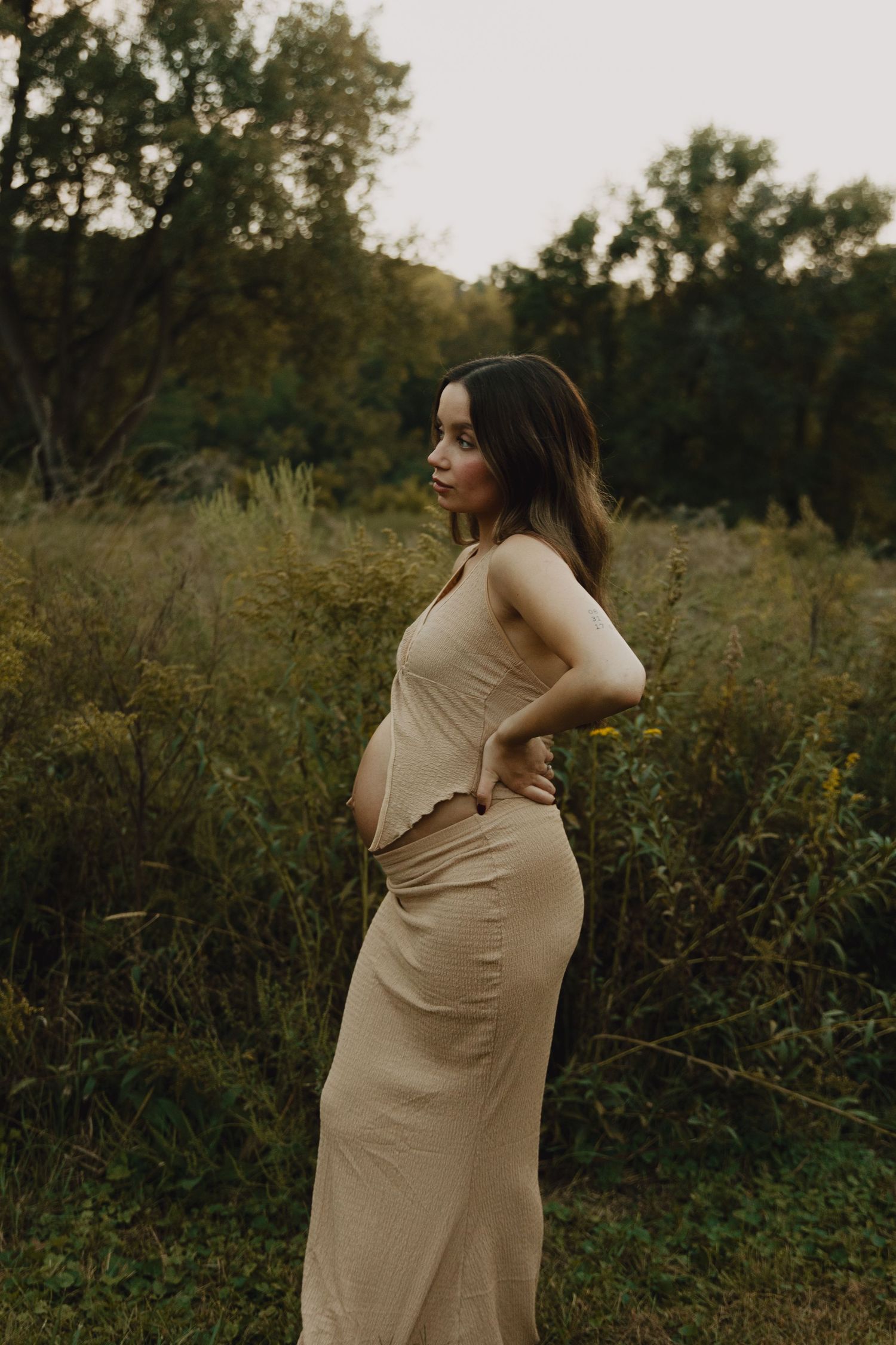 Side profile of expectant mother in a flowing beige dress standing in a natural outdoor setting.