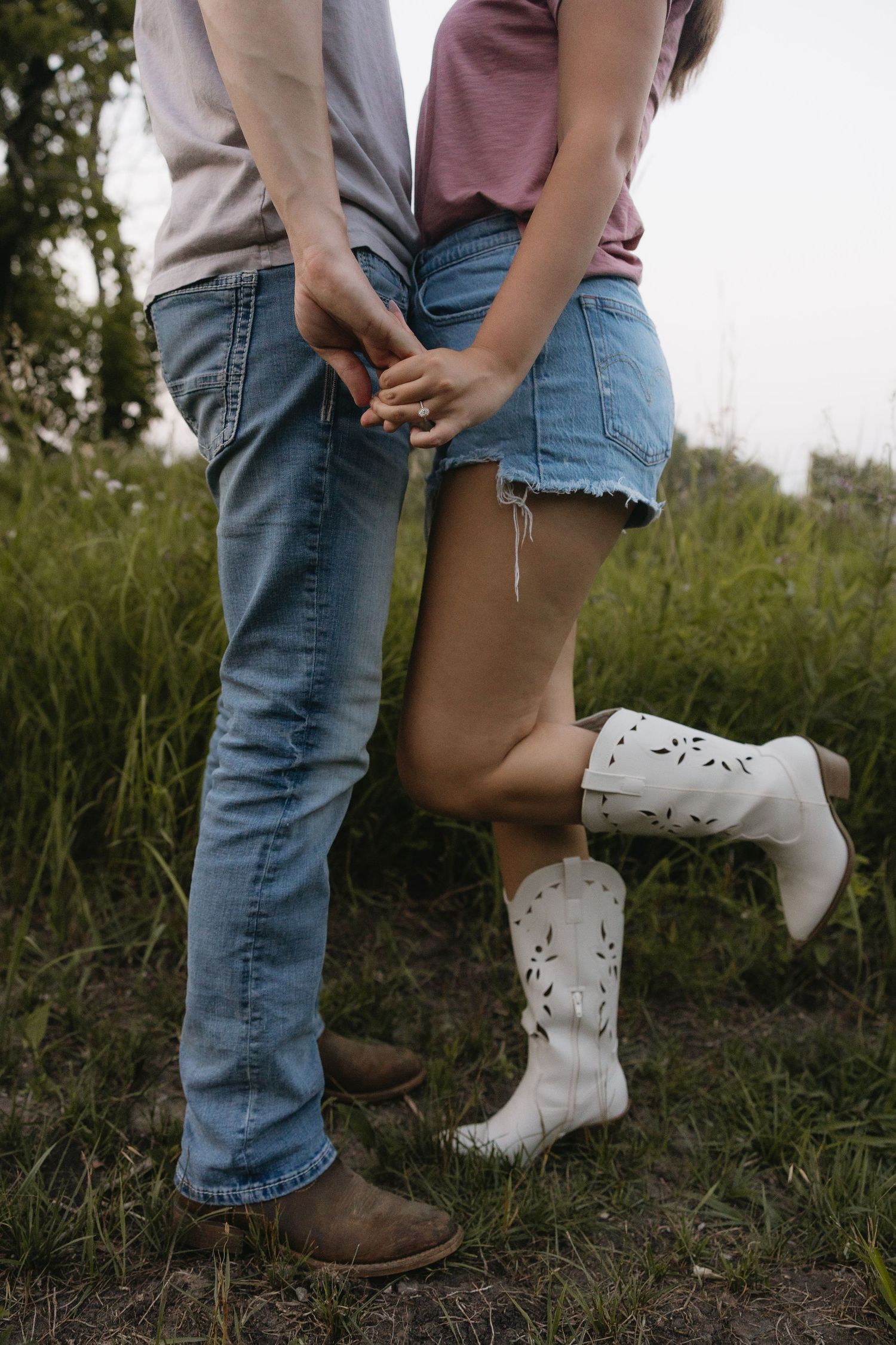 Couple holding hands in a field, wearing jeans and cowboy boots, with the woman in white western boots and denim shorts.