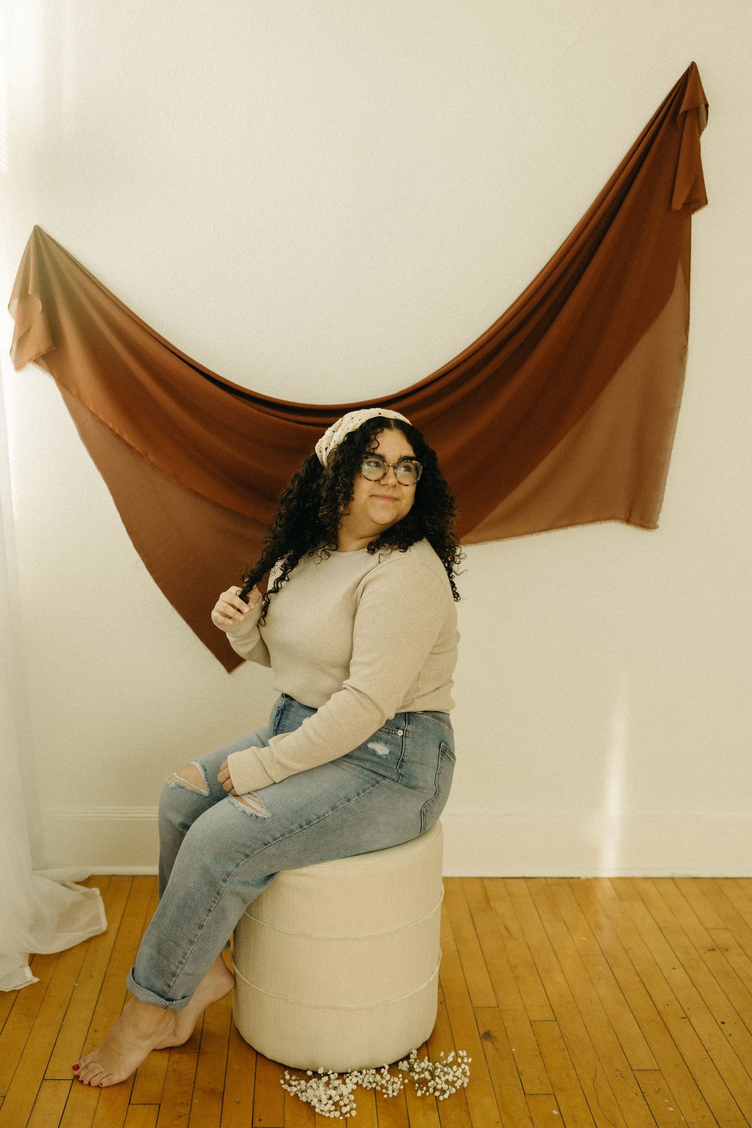 Woman with curly hair and glasses sits on a stool before a brown draped backdrop on a wood floor.