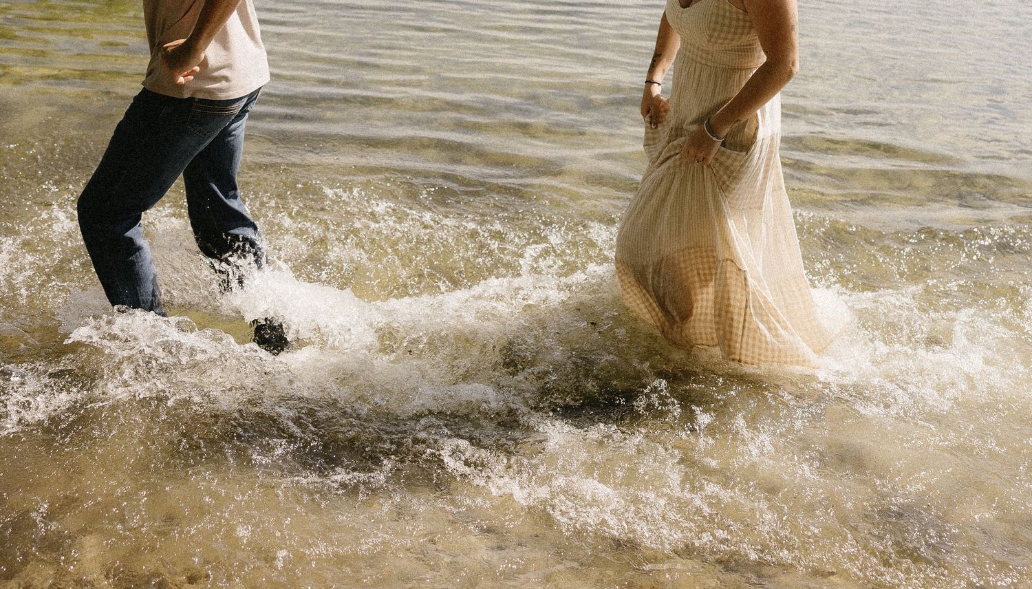 Couple wading through ocean waves at beach, woman in white wedding dress and man in casual attire.