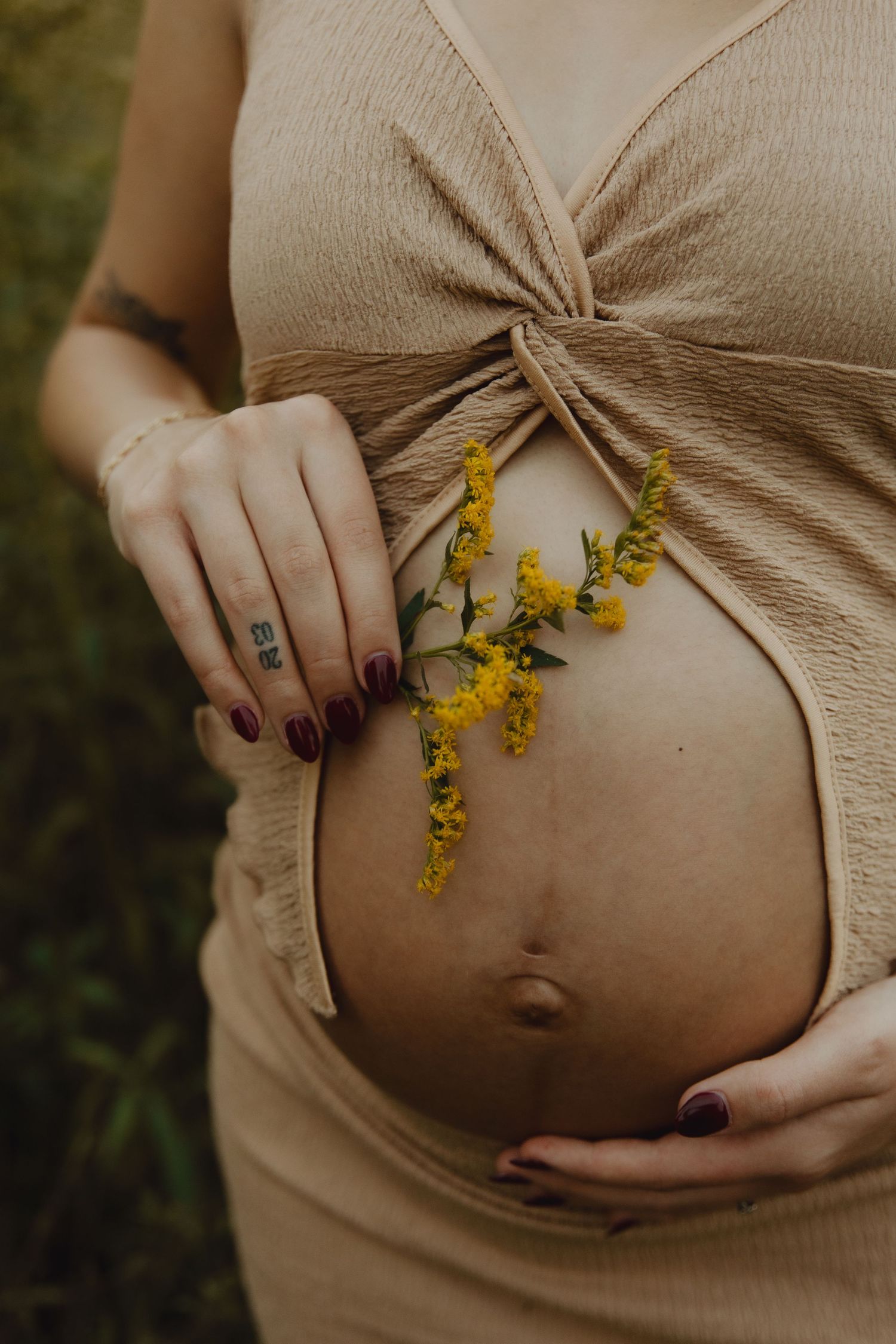 Close-up of pregnant belly with yellow wildflowers and purple nail polish in a beige maternity dress.