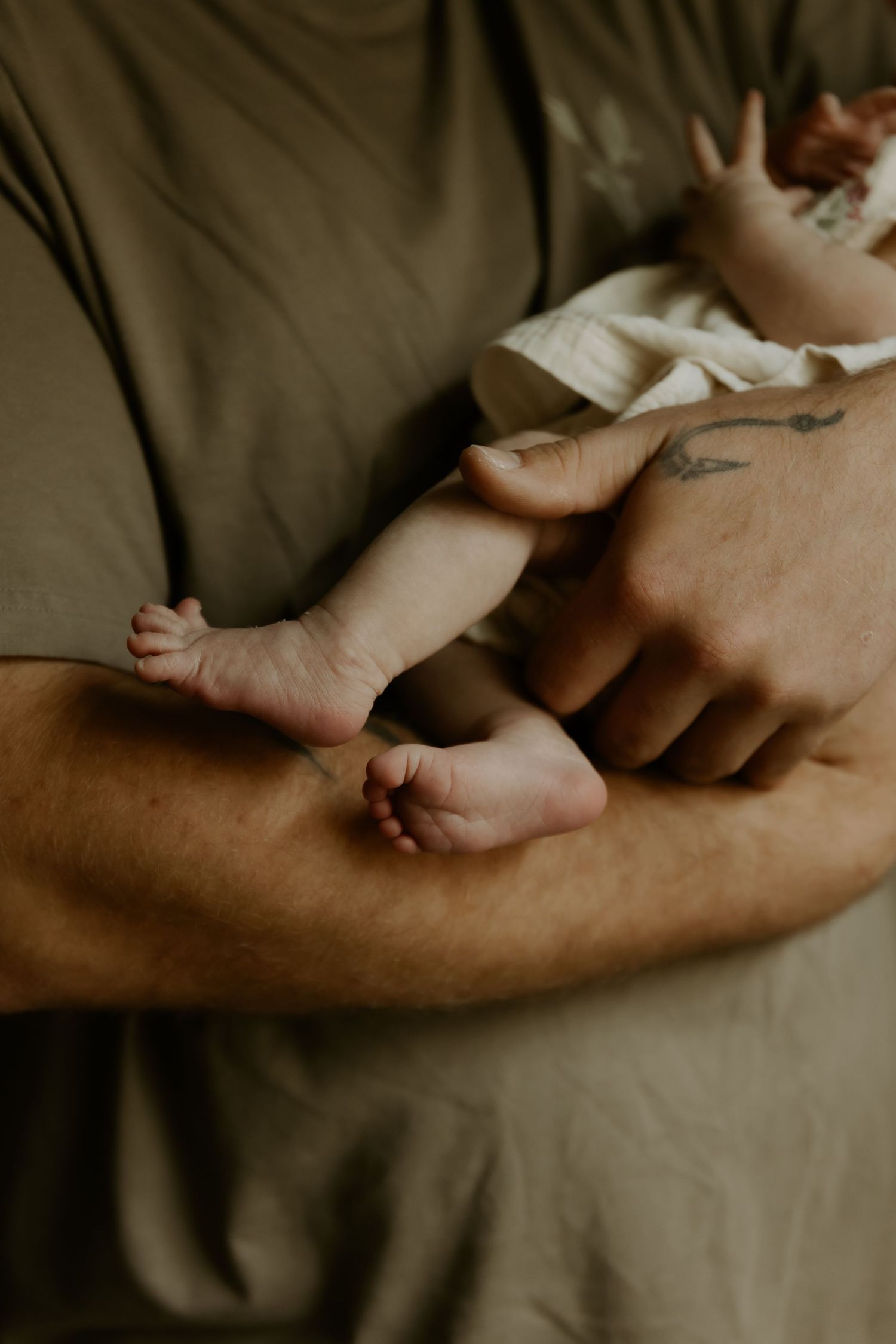 Artistic close-up of tiny baby feet resting against an adult's arm in muted lighting.