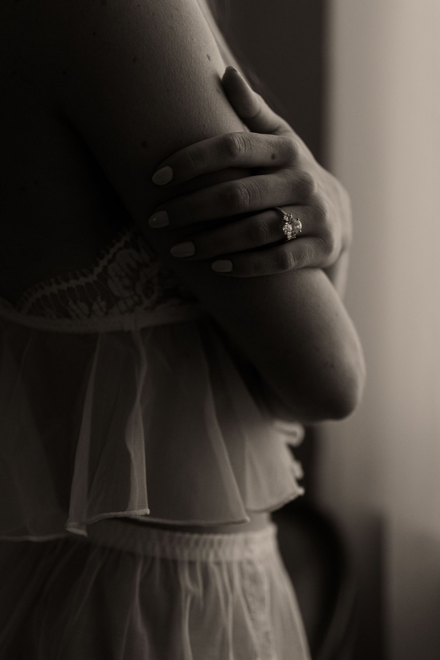 A close-up black and white photo showing hands with an engagement ring against a white dress.