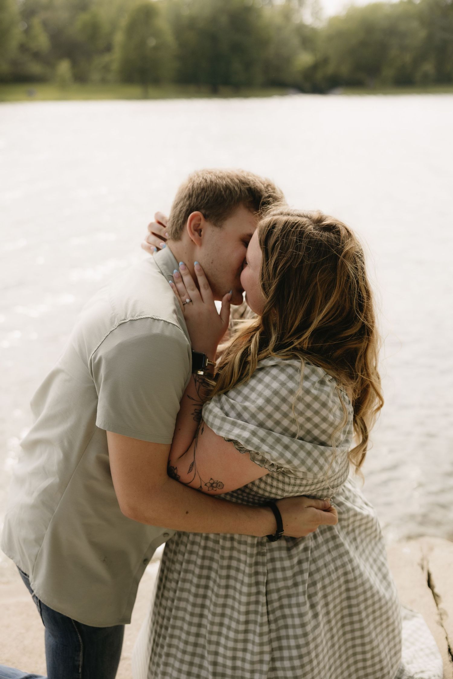 Couple sharing a romantic kiss by a lakeside, woman in gingham dress with tattoos embracing man in light shirt.