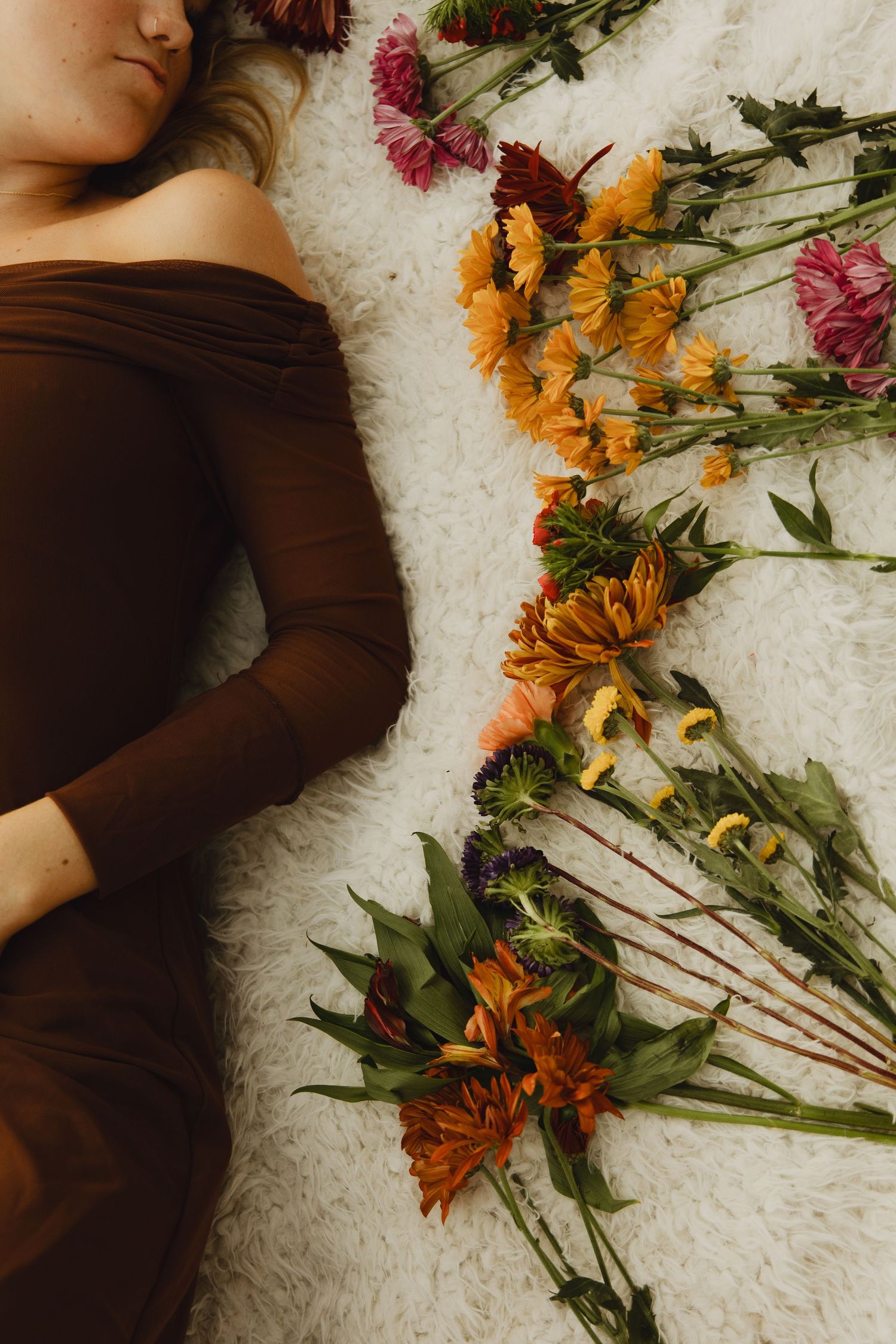 A person in brown clothing lies on white carpet beside scattered colorful autumn flowers and stems.